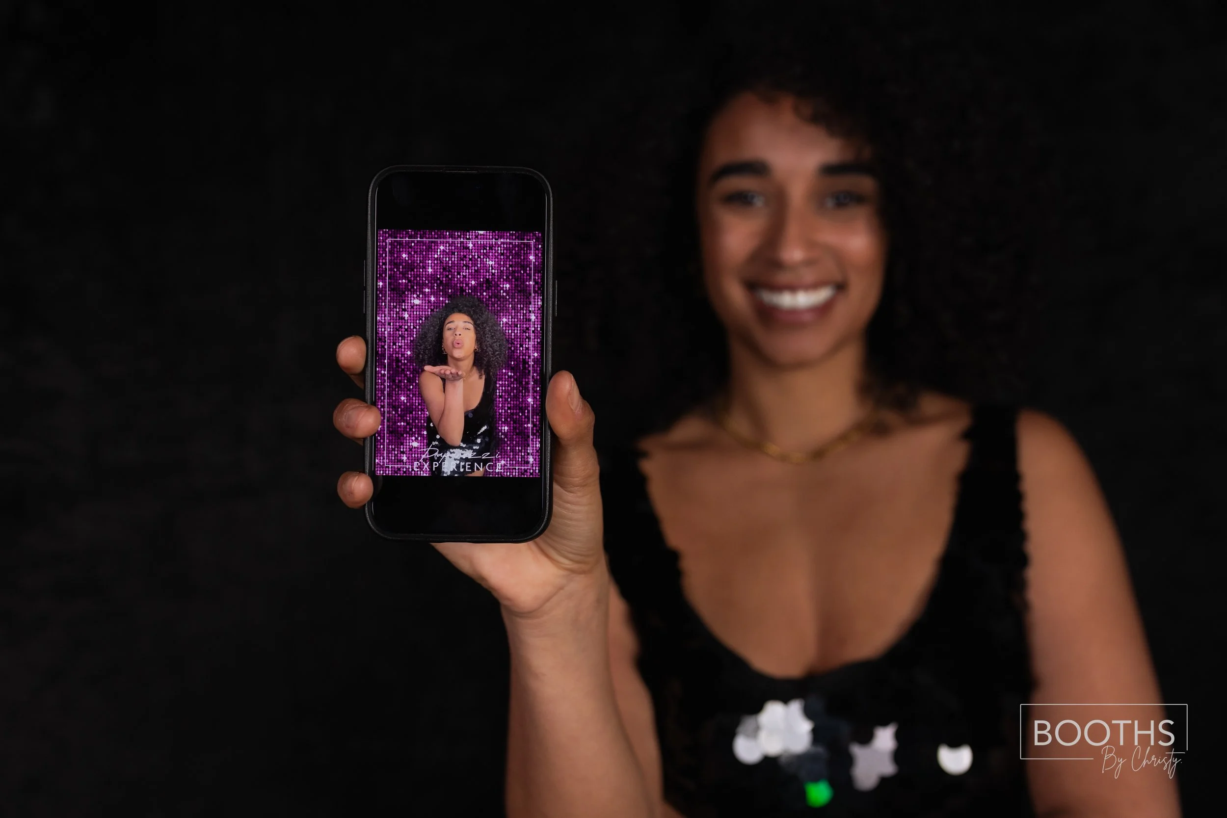 Woman with curly hair taking a selfie against a sparkly pink background, smiling, wearing a black top with sequin accents.