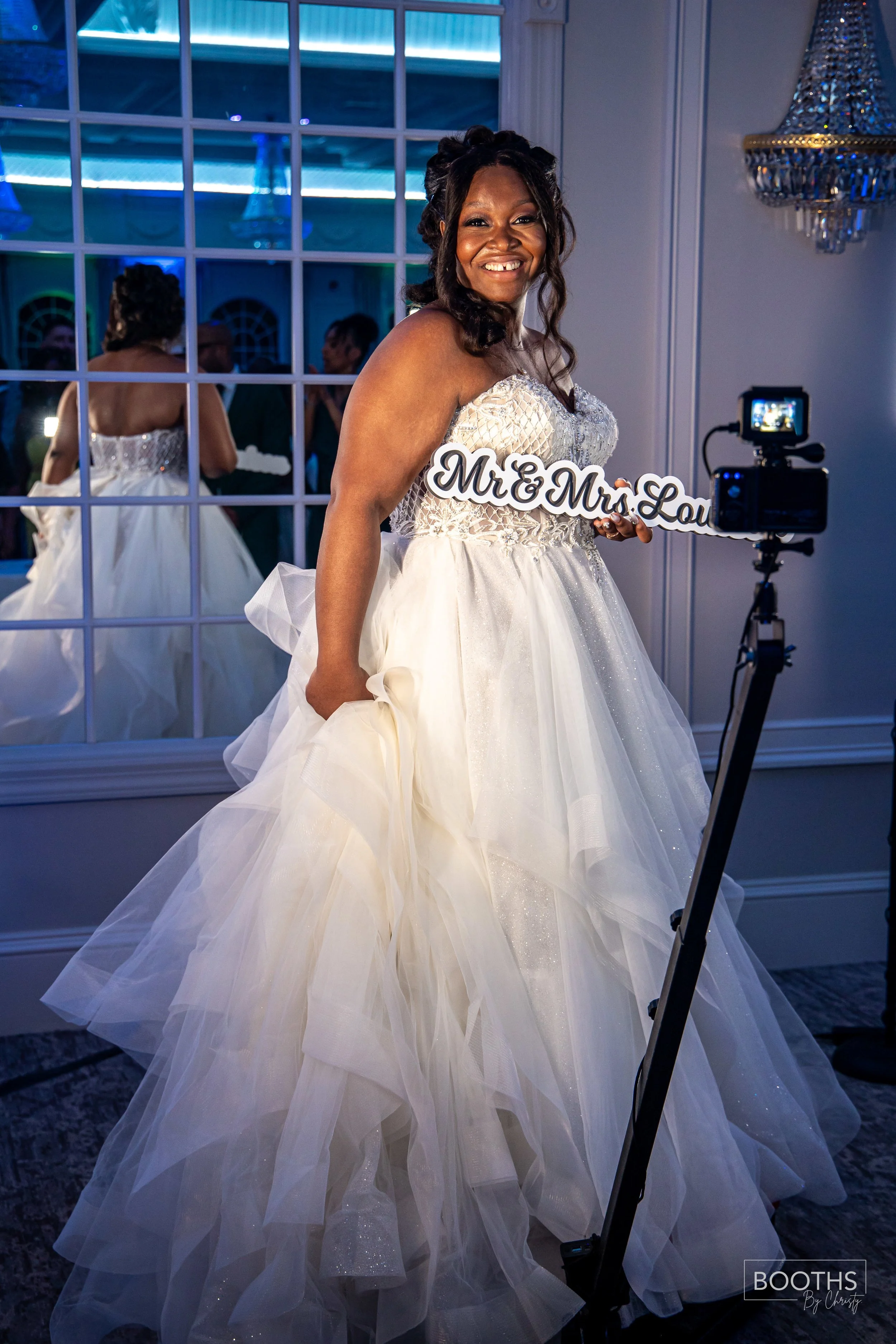 A bride in a white wedding gown holding a sign that says "Mr. & Mrs. Lou" at her wedding reception, standing in front of a mirror with a reflection of her back visible, in a decorated indoor venue with a chandelier.