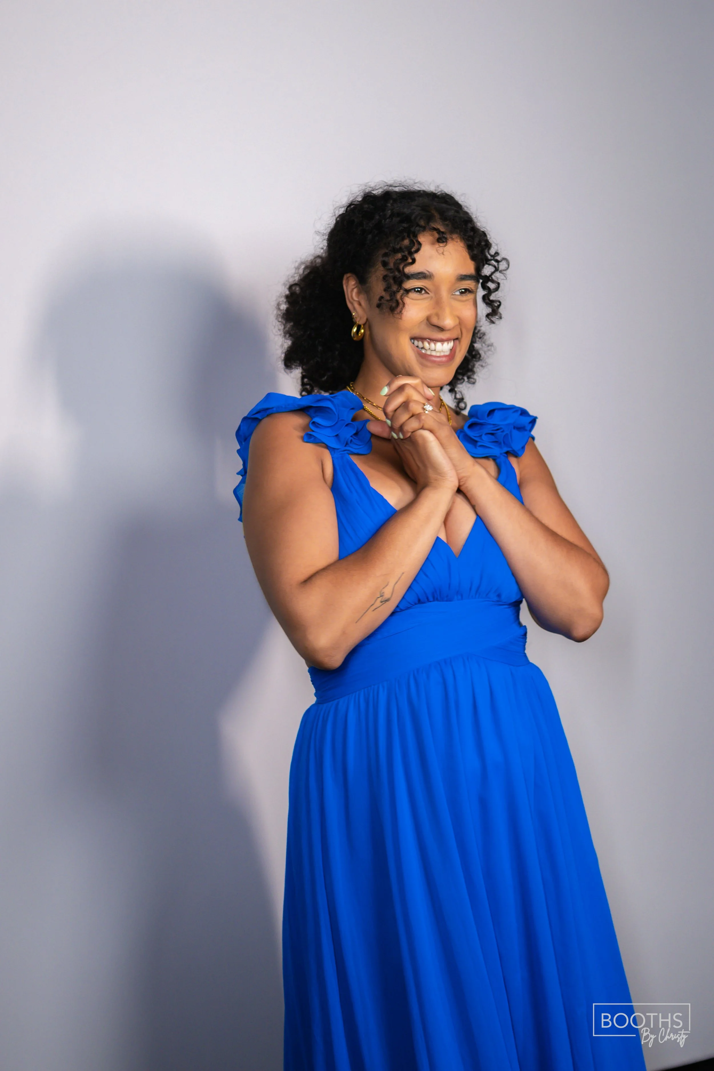 A woman with curly black hair wearing a bright blue dress with ruffled shoulders, smiling with her hands clasped near her chin, standing against a plain light gray background.