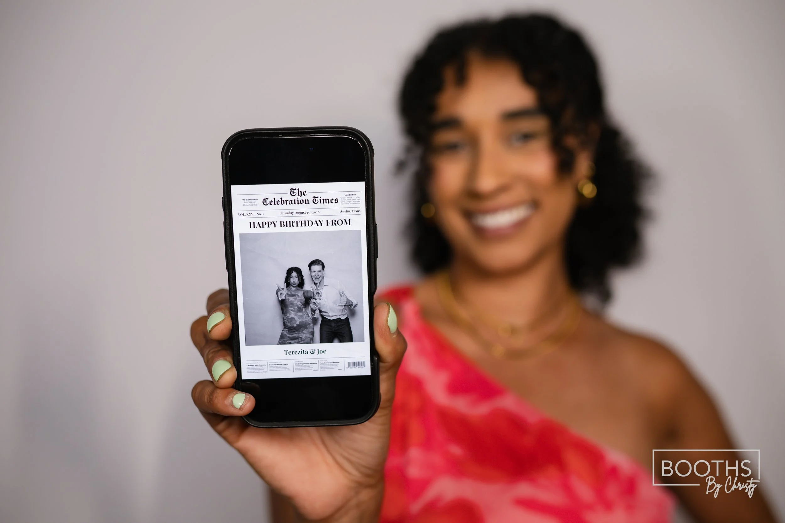 A woman smiling and holding a smartphone displaying a photo of two people with a newspaper headline that reads 'HAPPY BIRTHDAY FROM Terezia & Joe'.