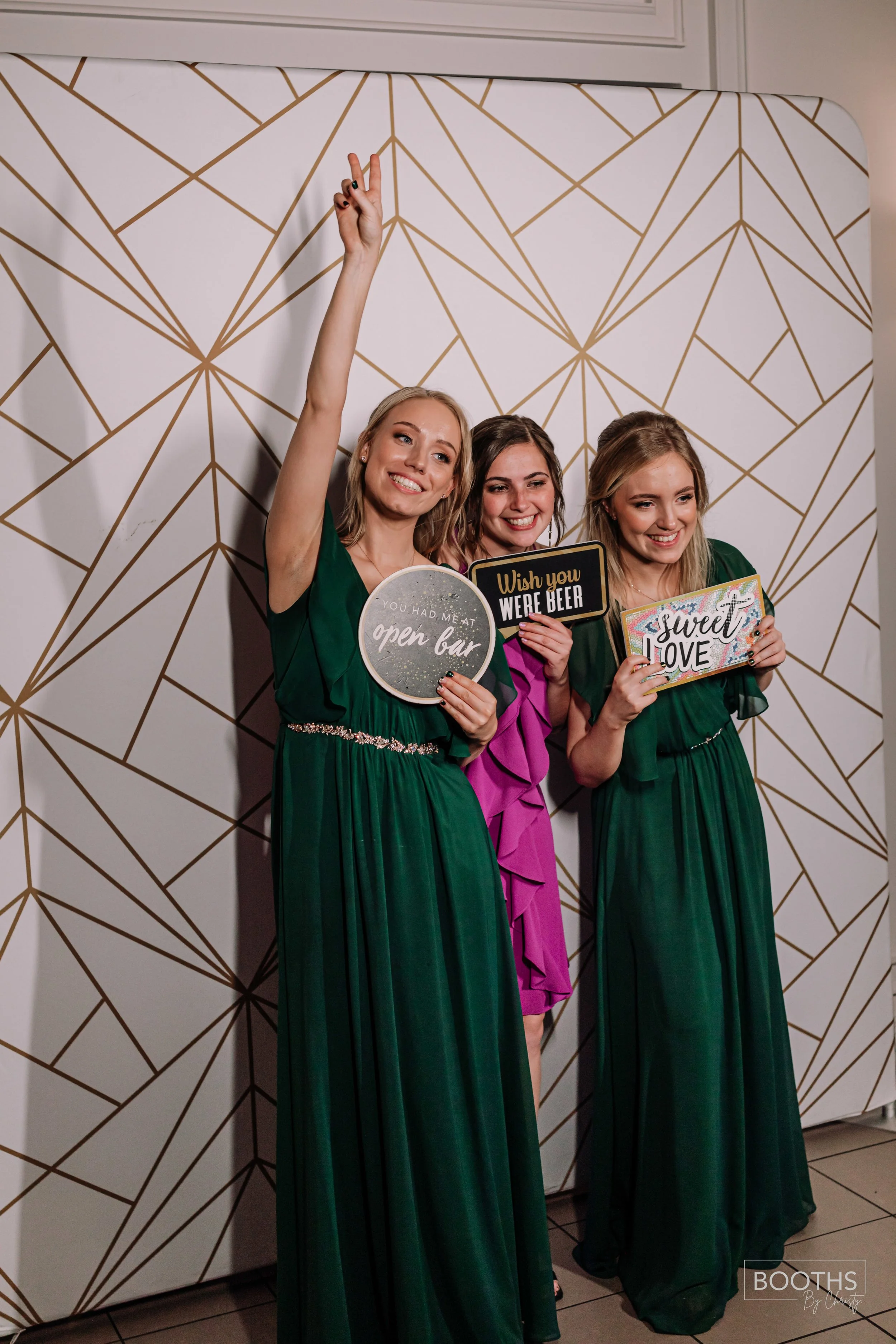 Three women in green and purple dresses holding humorous signs, smiling at a photo booth, with a geometric gold and white backdrop.