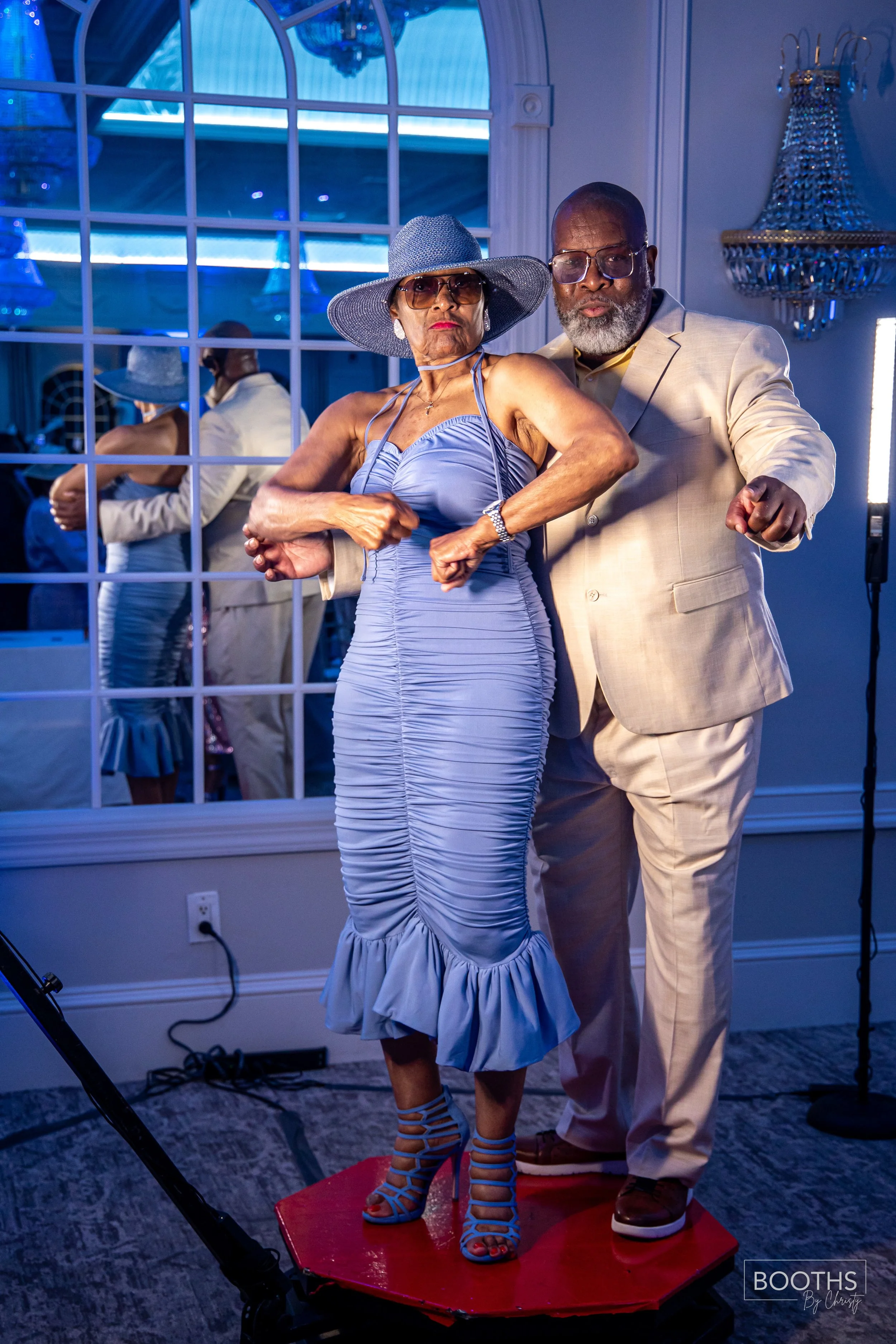 An elderly couple dancing on a small red platform at a party, with the woman wearing a blue dress, a wide-brimmed hat, and high heels, and the man in a light-colored suit. They are in a room with a mirror and a chandelier, dancing enthusiastically.