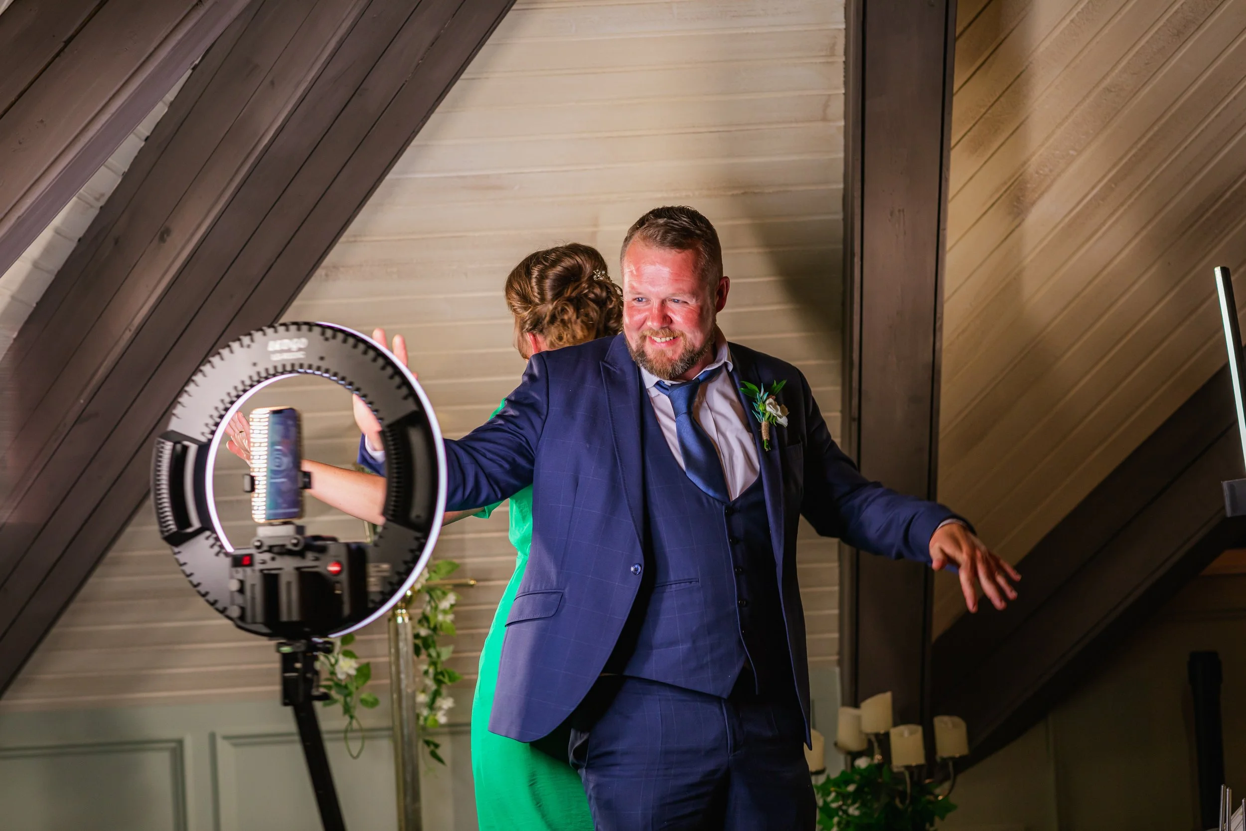 A man in a blue suit dancing during a celebration, with a ring light and a woman in a green dress partly visible behind him, in an indoor venue with wooden walls and candles.
