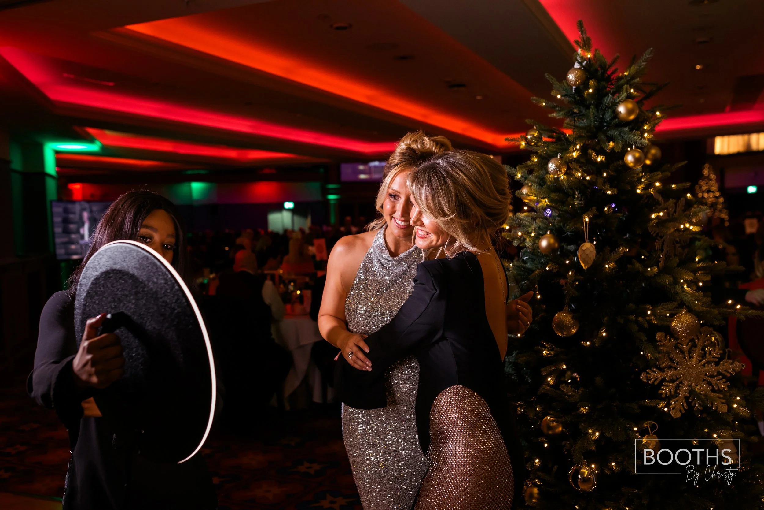 Three women in formal attire enjoy dancing near a decorated Christmas tree in a festive setting with colorful lighting.