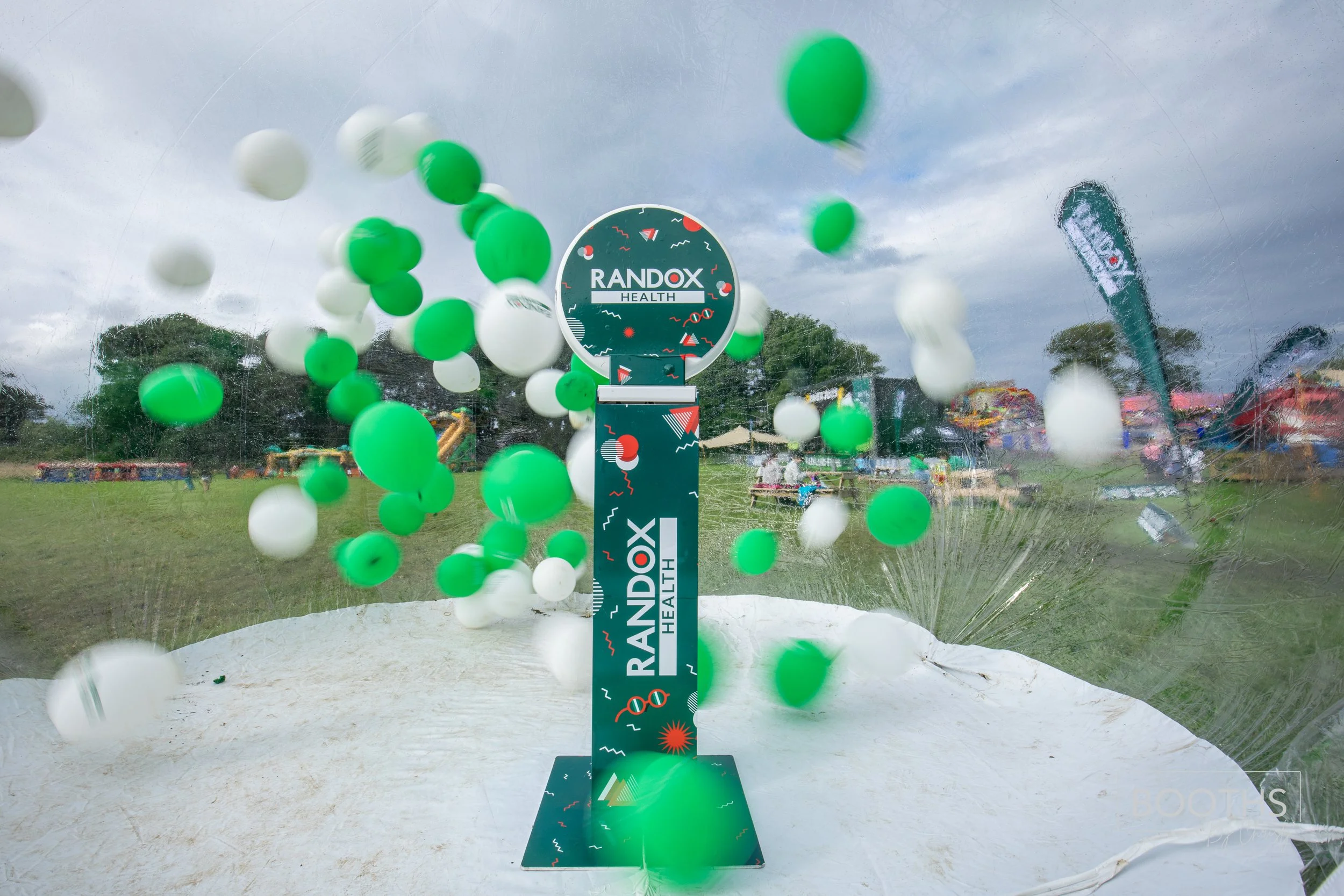 A display of green and white balloons bursting at an event with a sign that reads 'RANDOX HEALTH', outdoors on a cloudy day.