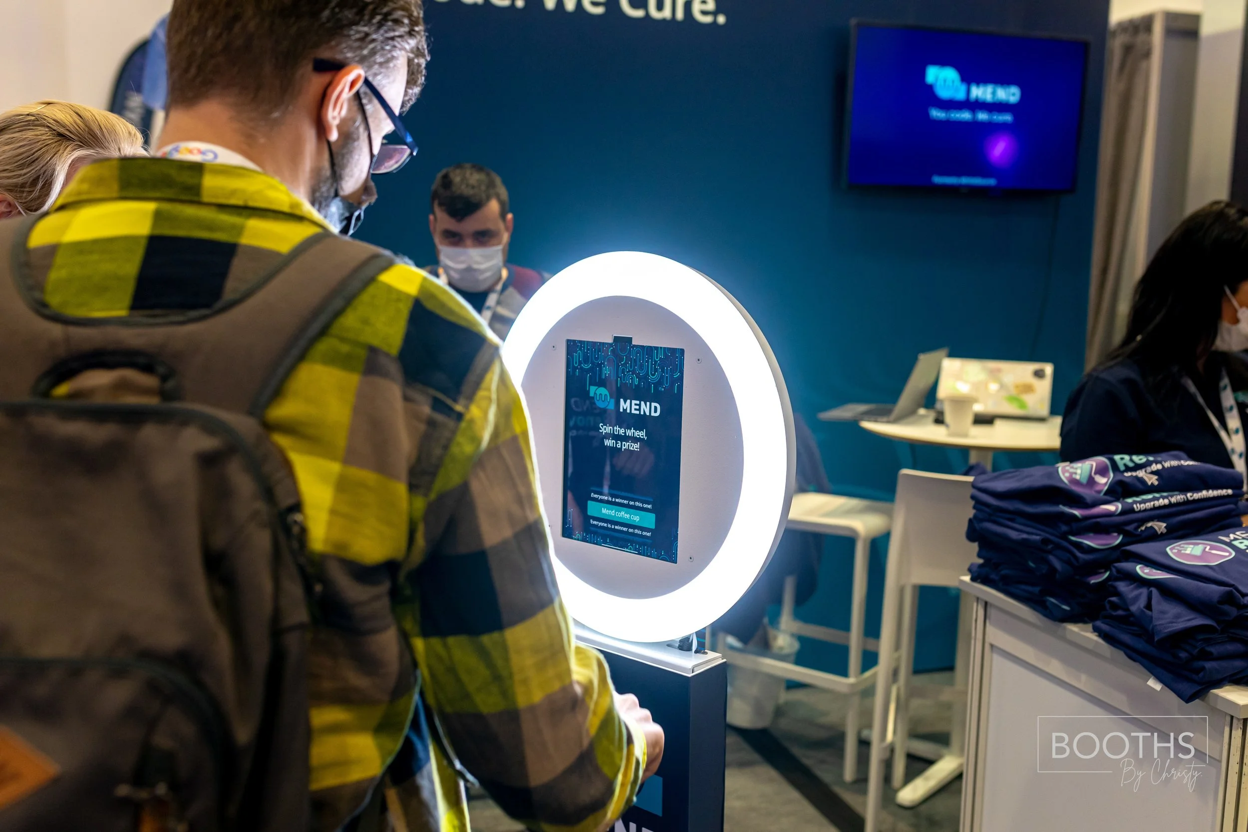 Person in a yellow and black checkered shirt and backpack using a circular digital kiosk with a blue screen inside a conference or event room. Several people wearing masks are in the background, and a display screen on the wall shows a blue screen.