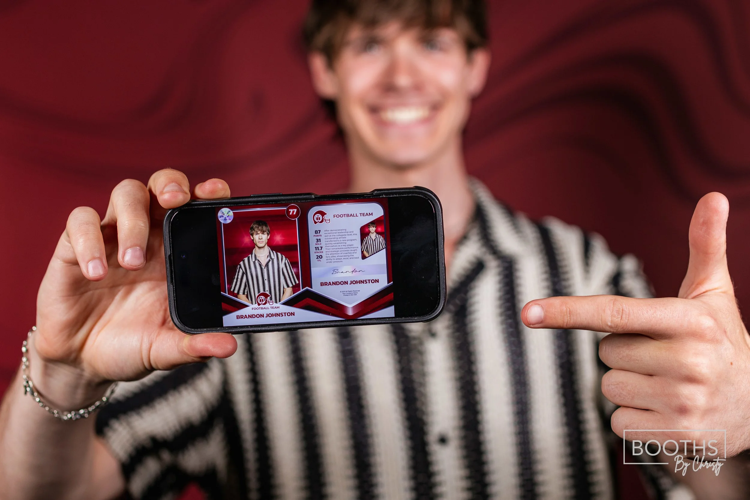 Person holding a smartphone displaying a football trading card, smiling, and pointing at it with their other hand, wearing a black and white striped shirt.