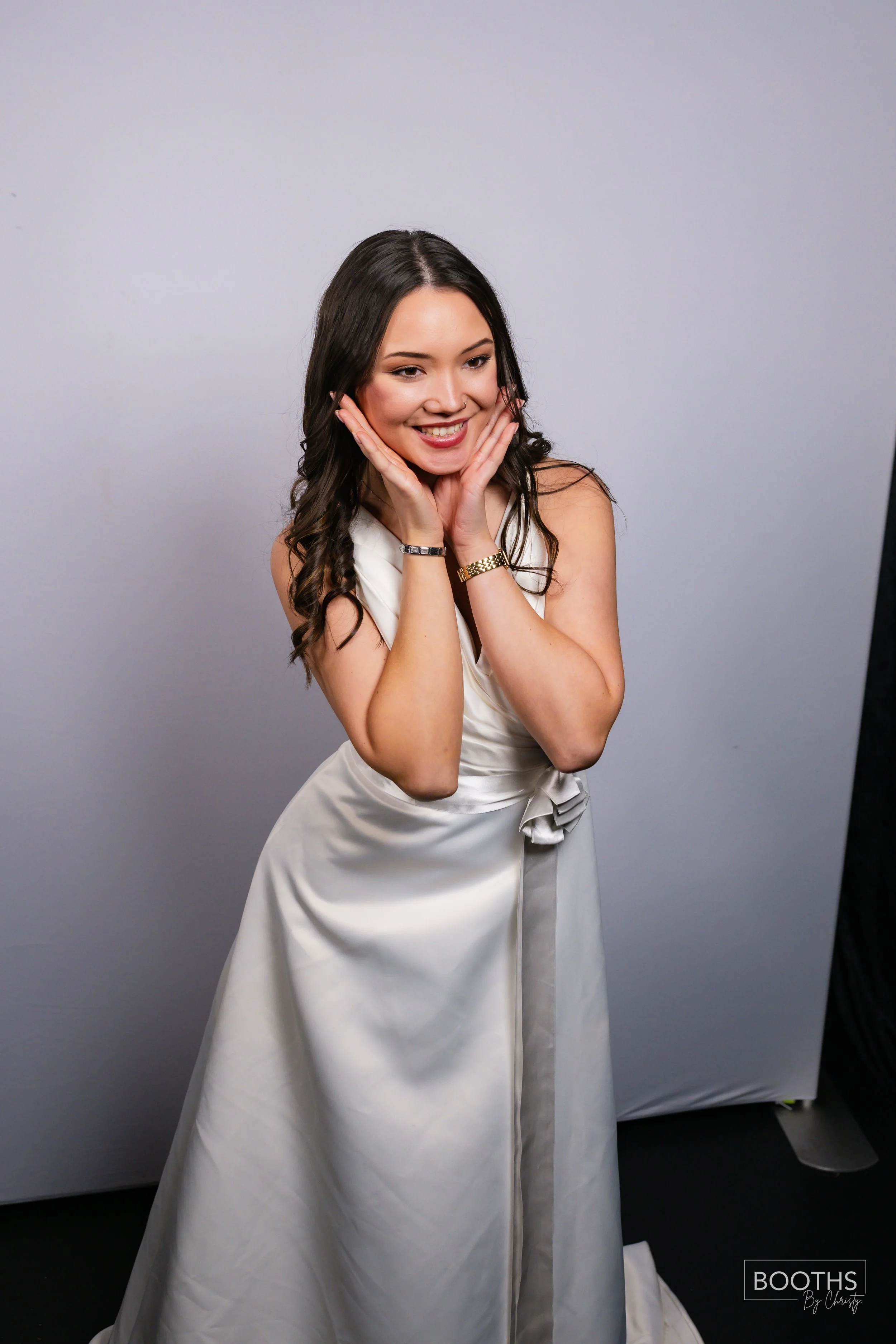 A young woman with long dark hair wearing a white satin dress with a bow, smiling and holding her face with both hands, standing against a plain grey background.