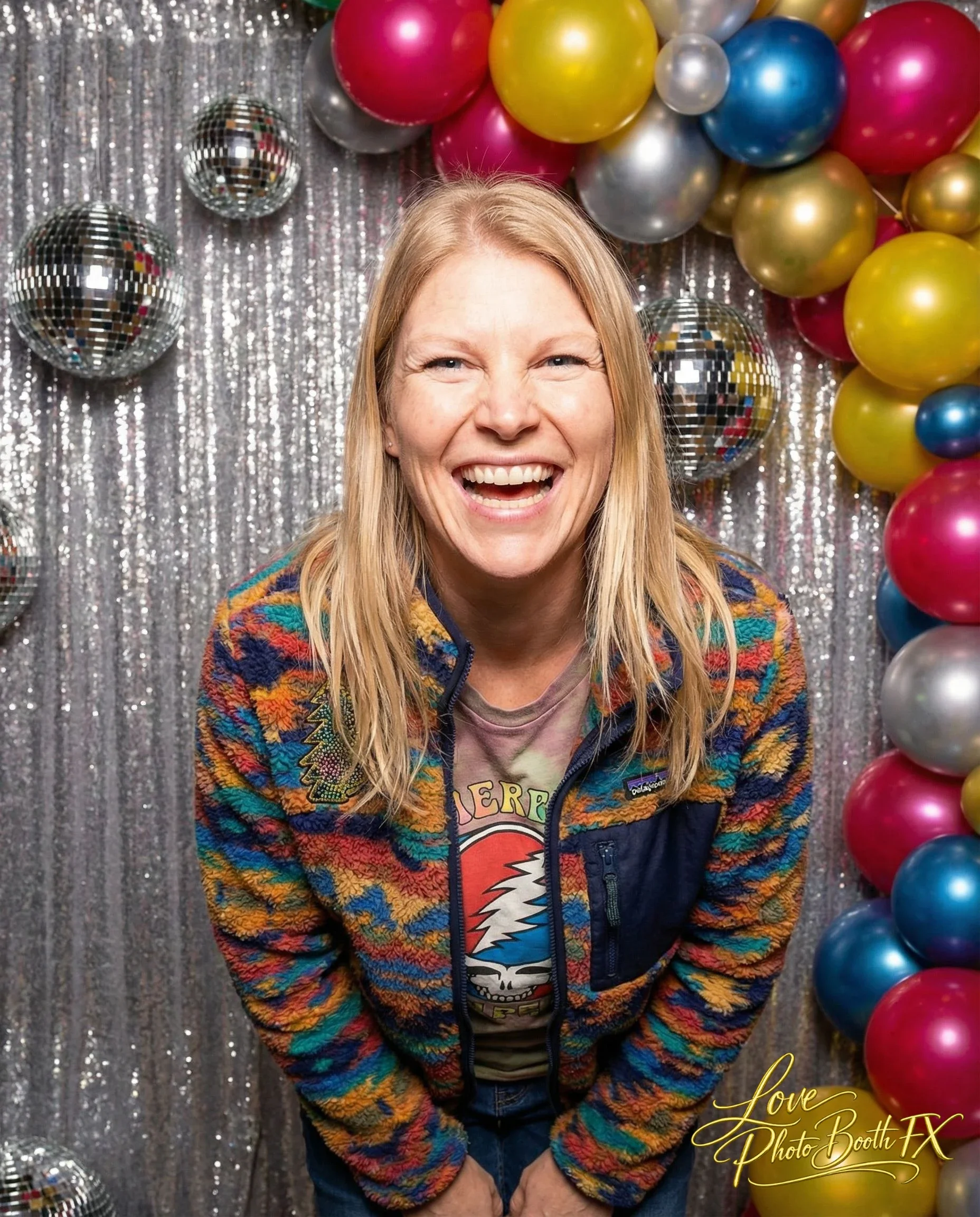 A woman with long blonde hair smiling and laughing, wearing a colorful patterned jacket and a Grateful Dead t-shirt, standing in front of a silver sequin backdrop decorated with balloons and disco balls.