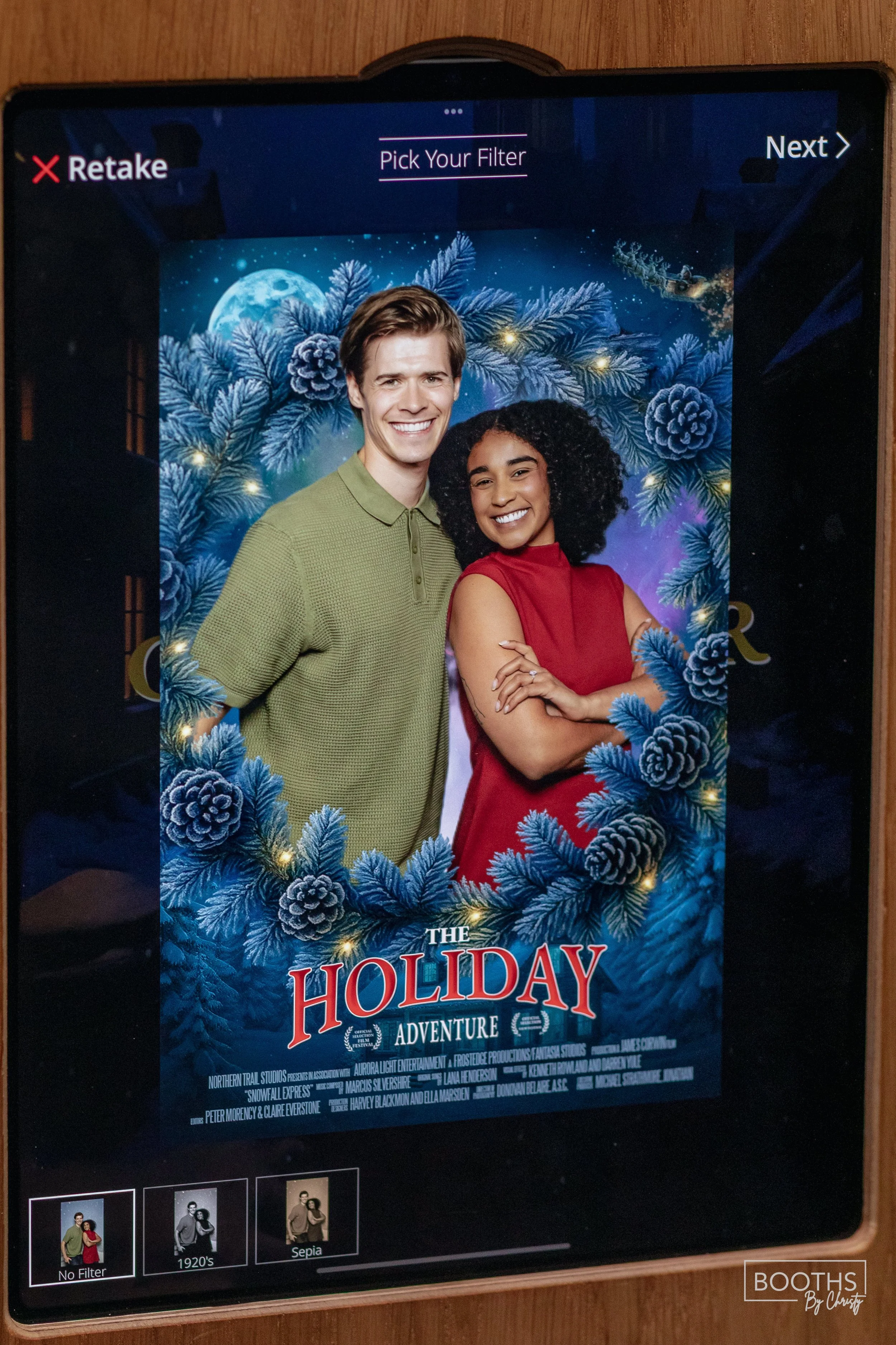 Digital poster for the holiday movie "The Holiday Adventure" featuring a smiling young man in a green shirt and a young woman in a red dress, surrounded by a festive blue pine branch wreath with pinecones, set against a snowy night scene with a full
