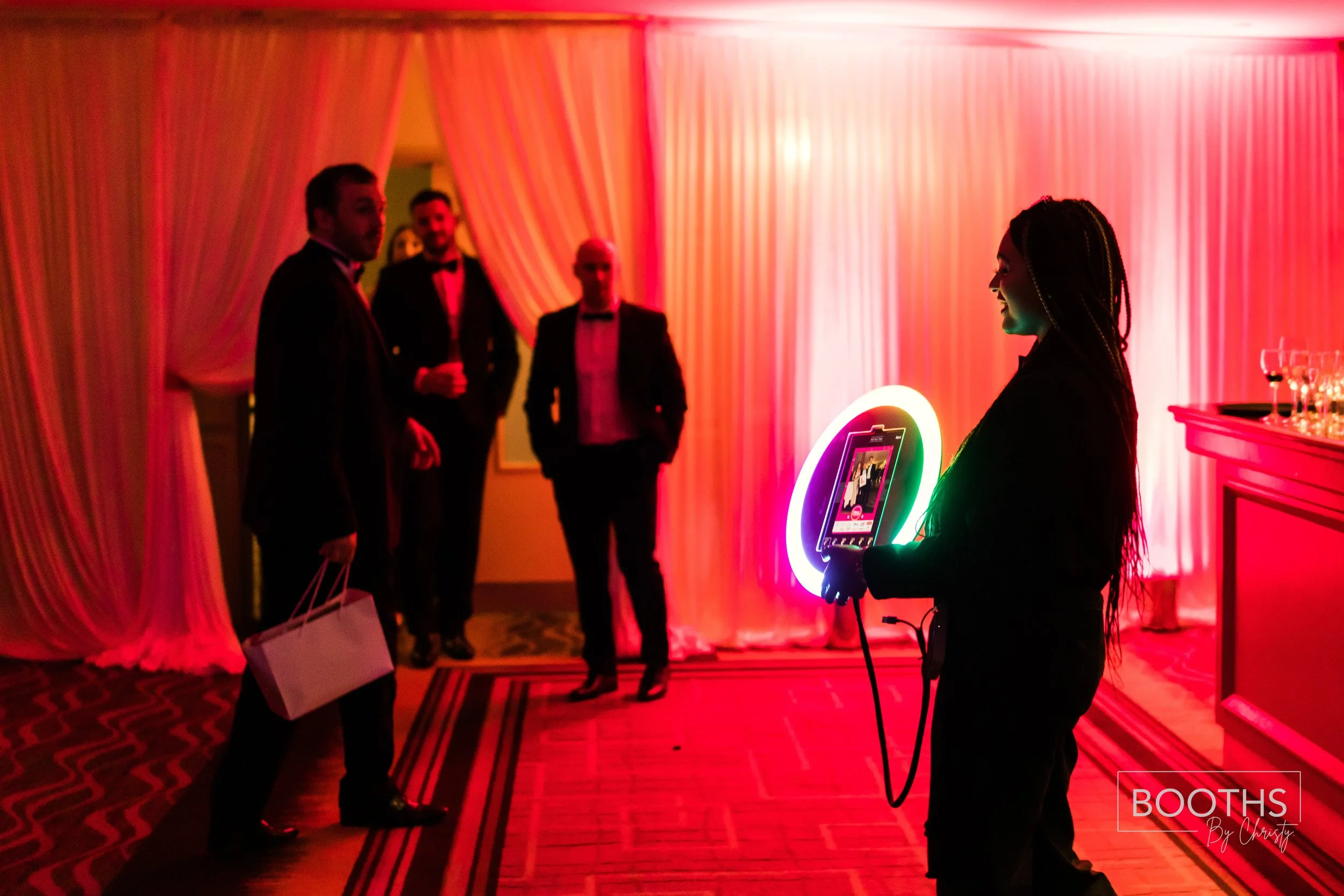 A group of people dressed in formal attire standing in a party setting with red and pink lighting, with a woman holding a neon-lit tablet or photo booth device.