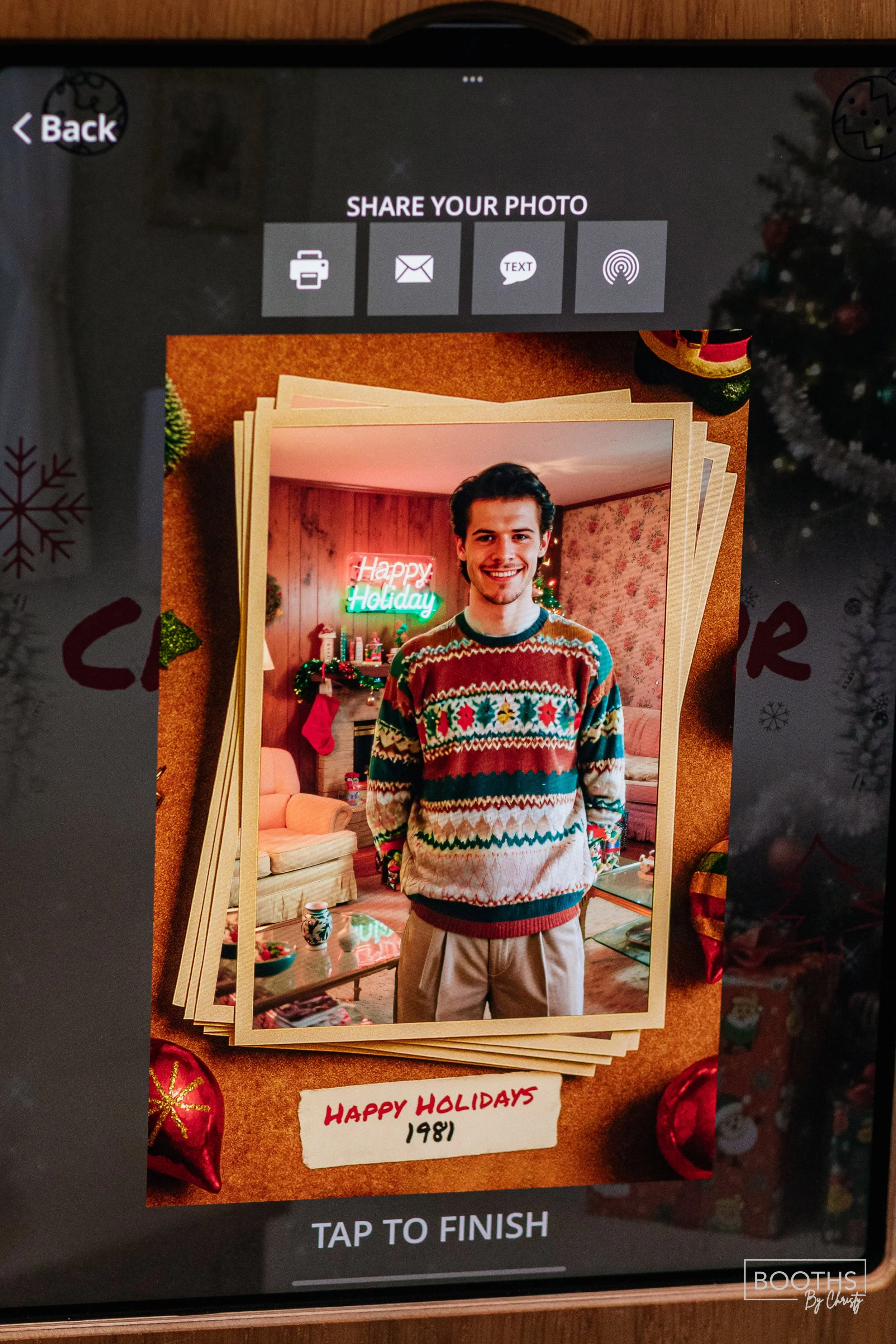 A young man with dark hair and a mustache is smiling and standing in a decorated Christmas room, wearing a colorful holiday sweater. Behind him, a neon sign says "Happy Holiday" and the room is decorated with Christmas stockings, ornaments, and garla