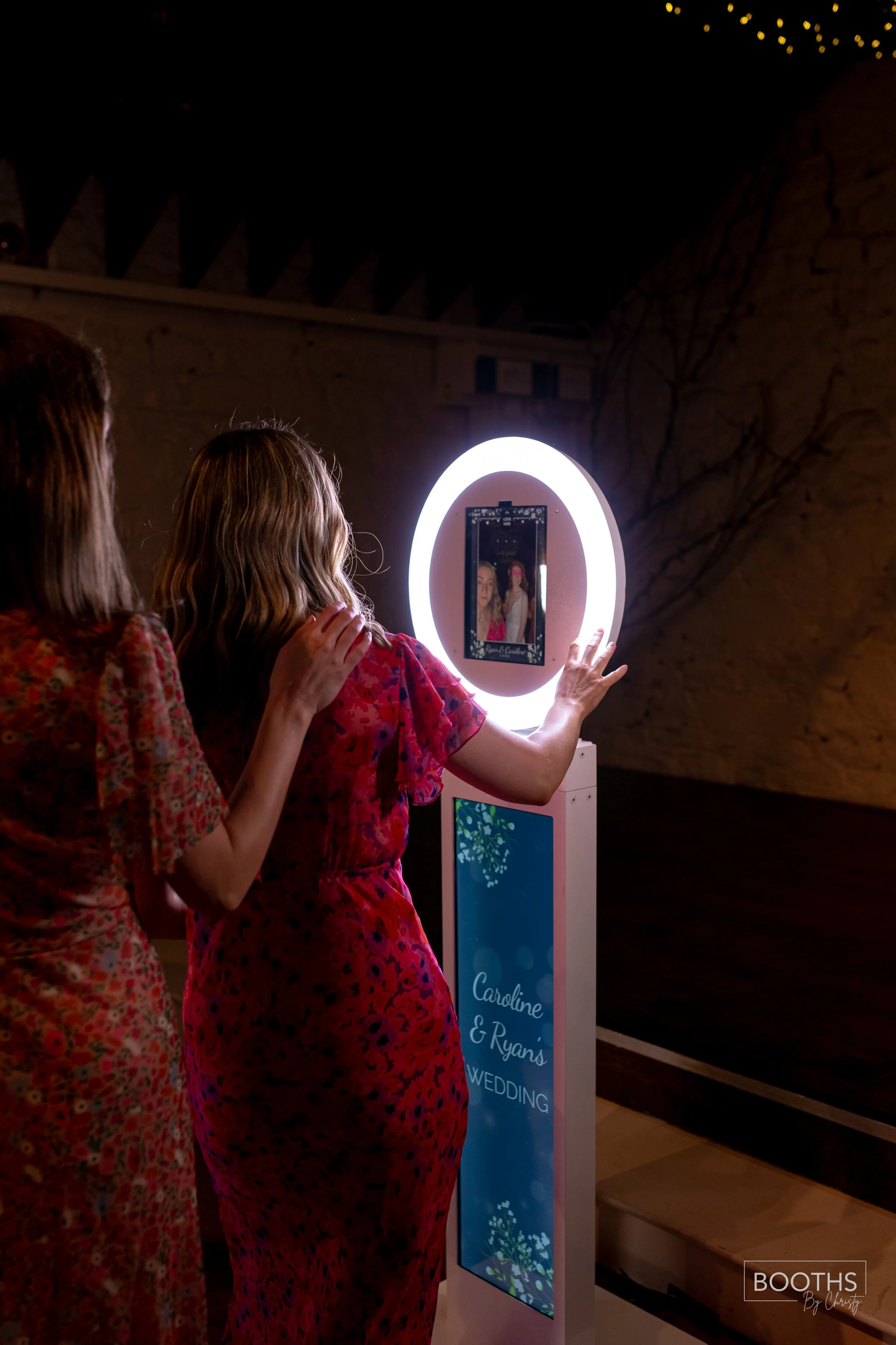 A woman and a girl using a lit-up photo booth mirror at a wedding reception. The mirror displays an image of the two taking a selfie. The stand has a sign that reads 'Caroline & Ryan's Wedding'.