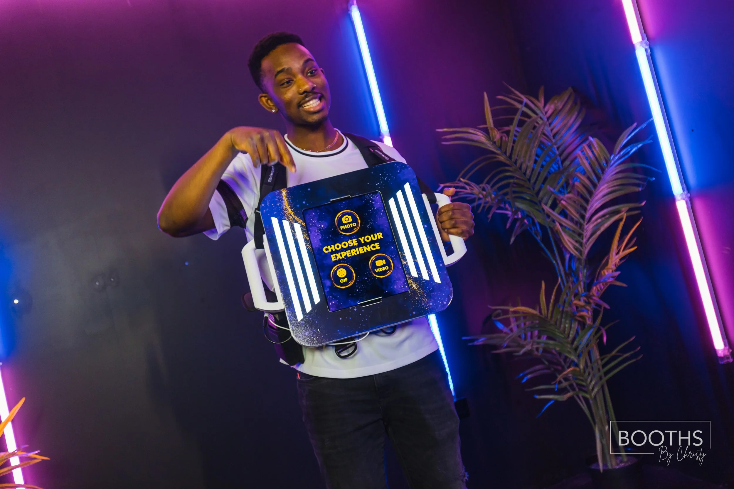 Young man with short hair, wearing a black backpack and a white t-shirt, smiling and pointing to a sign that says "Choose Your Experience" with icons for photo, selfie, GIF, and video. The background features dark walls with colorful neon lights and