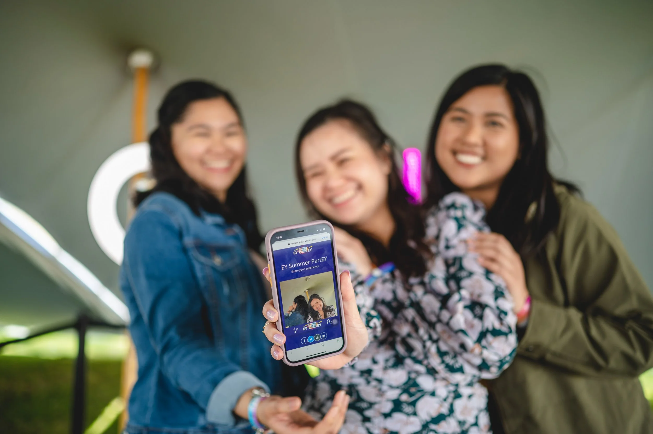 Three young women smiling and taking a selfie at an outdoor event, with one holding a phone displaying a photo from the event titled 'EY Summer Party'.