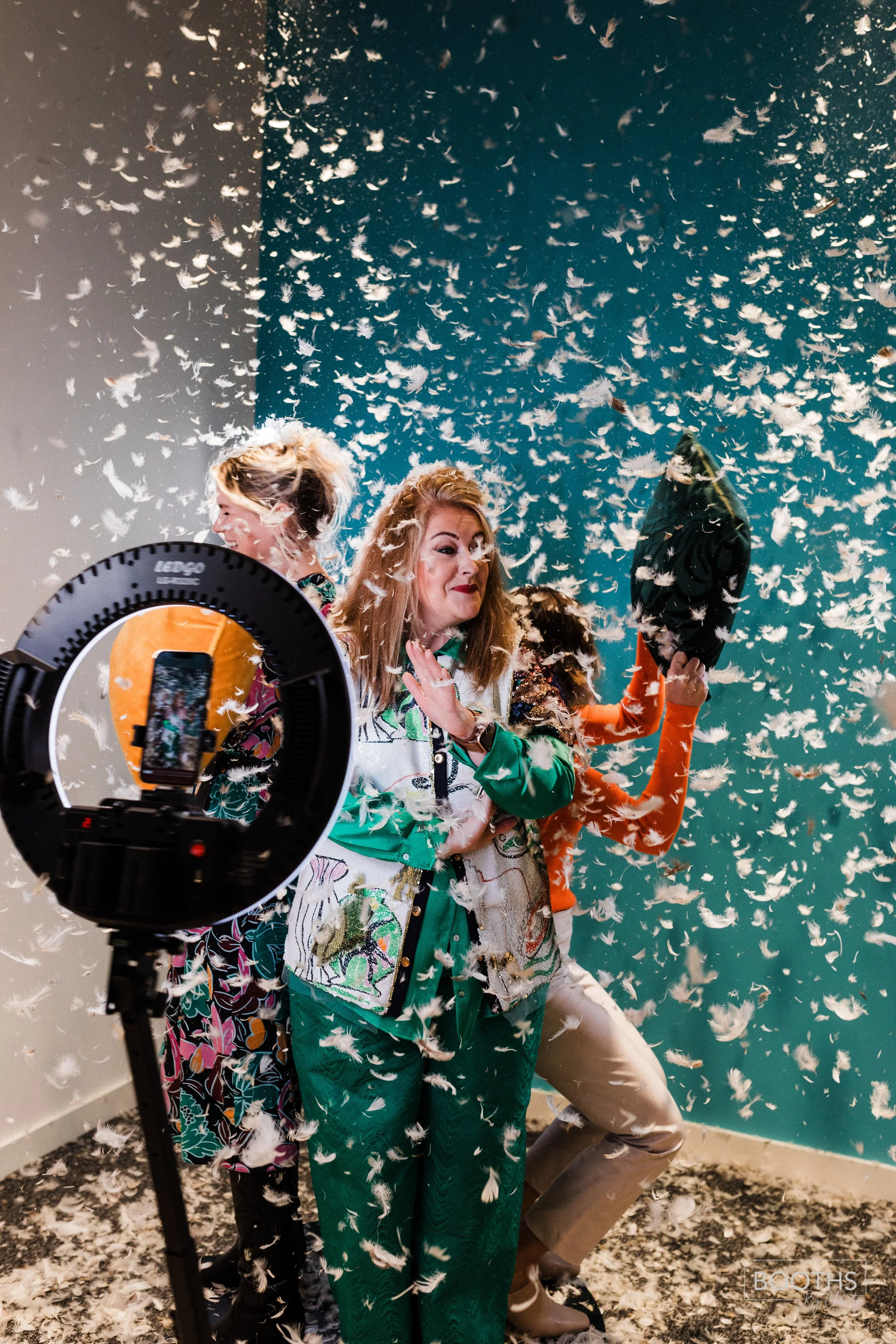 Three women standing in a room with blue walls, surrounded by falling white feathers. One woman in the center is holding a black hat and smiling, while the other two women are partially visible behind her.