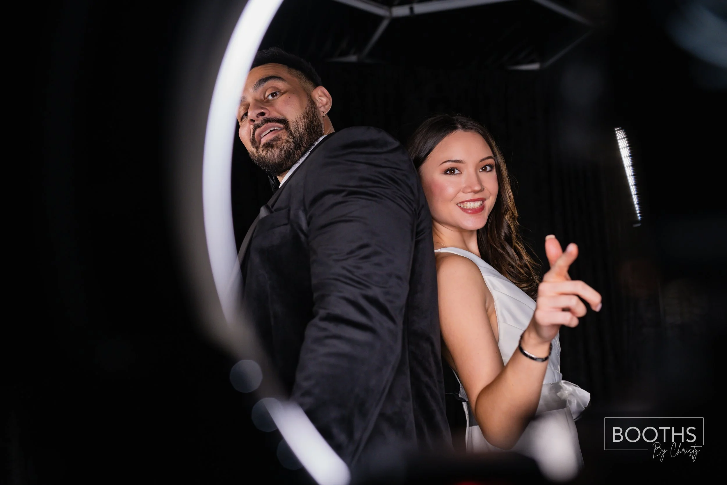 A man and woman dressed formal, standing back to back, smiling at the camera, with a dark background. The woman is making a finger gun gesture, and the photo is framed by a ring light.