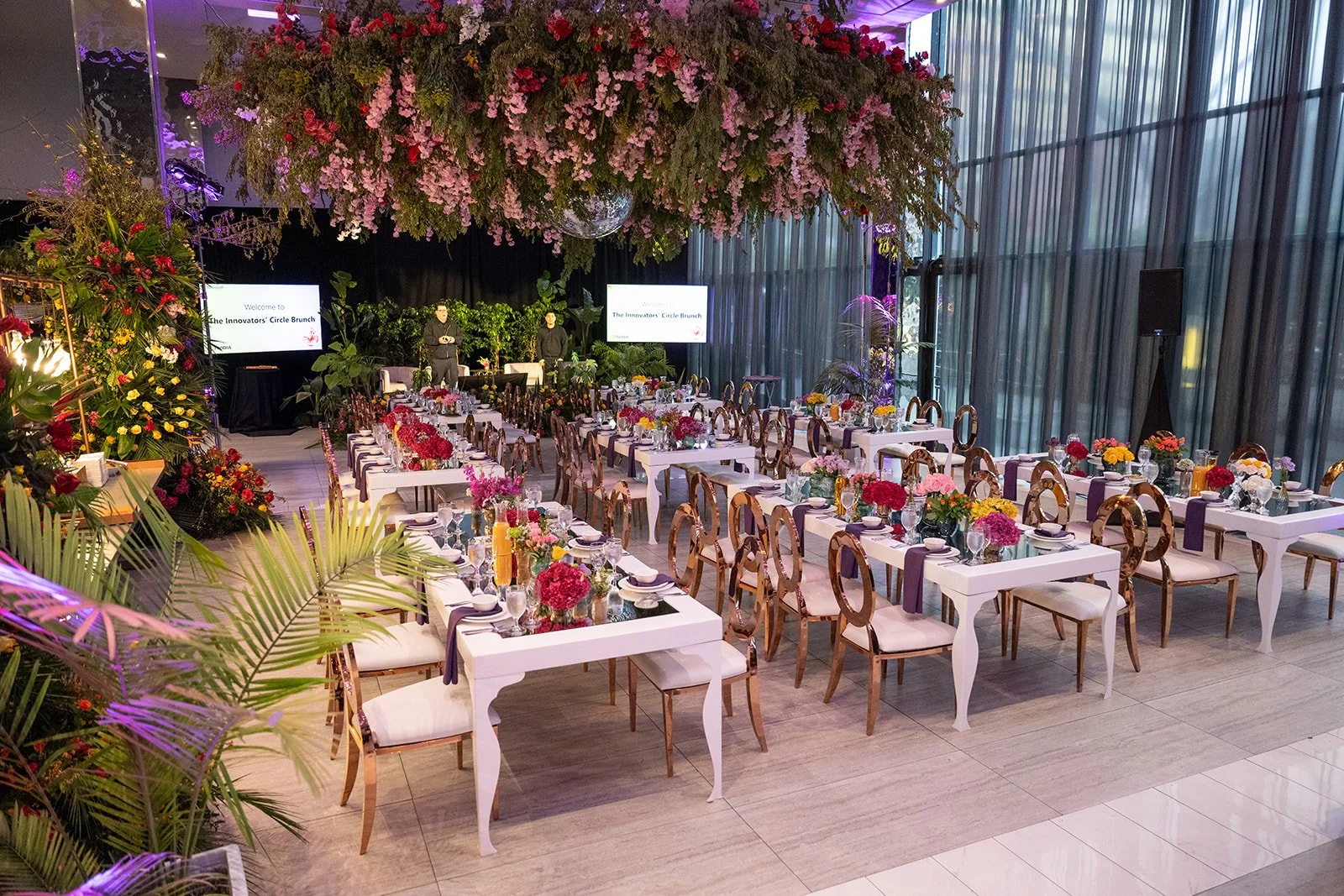 Elegant banquet hall decorated with floral arrangements and a large hanging flower installation, set for a formal event with tables, chairs, and presentation screens.