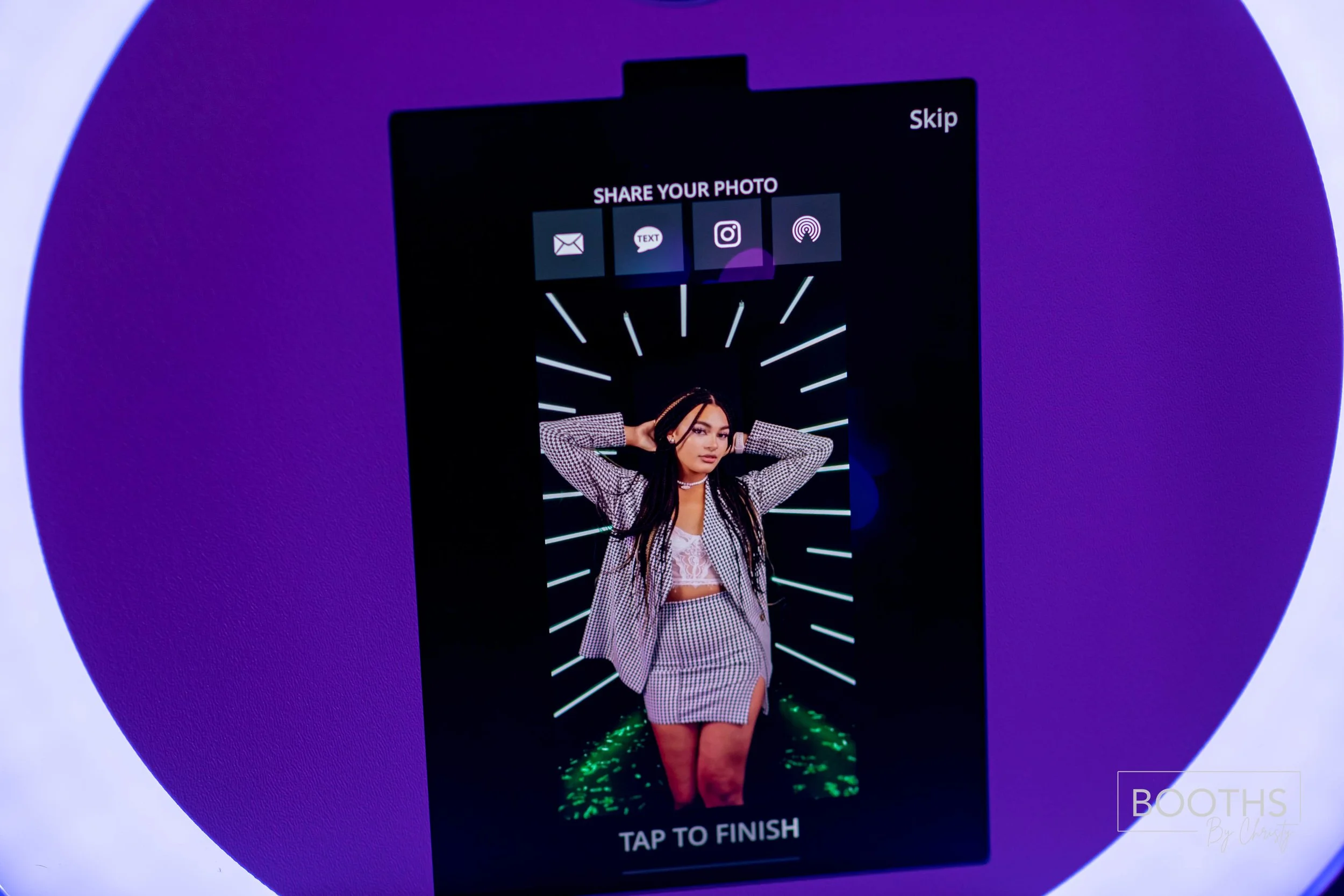 A girl taking a photo at a photo booth, wearing a checkered outfit and headphones, with a digital interface for sharing the picture above her head. The background features neon lights and the text "TAP TO FINISH" at the bottom.