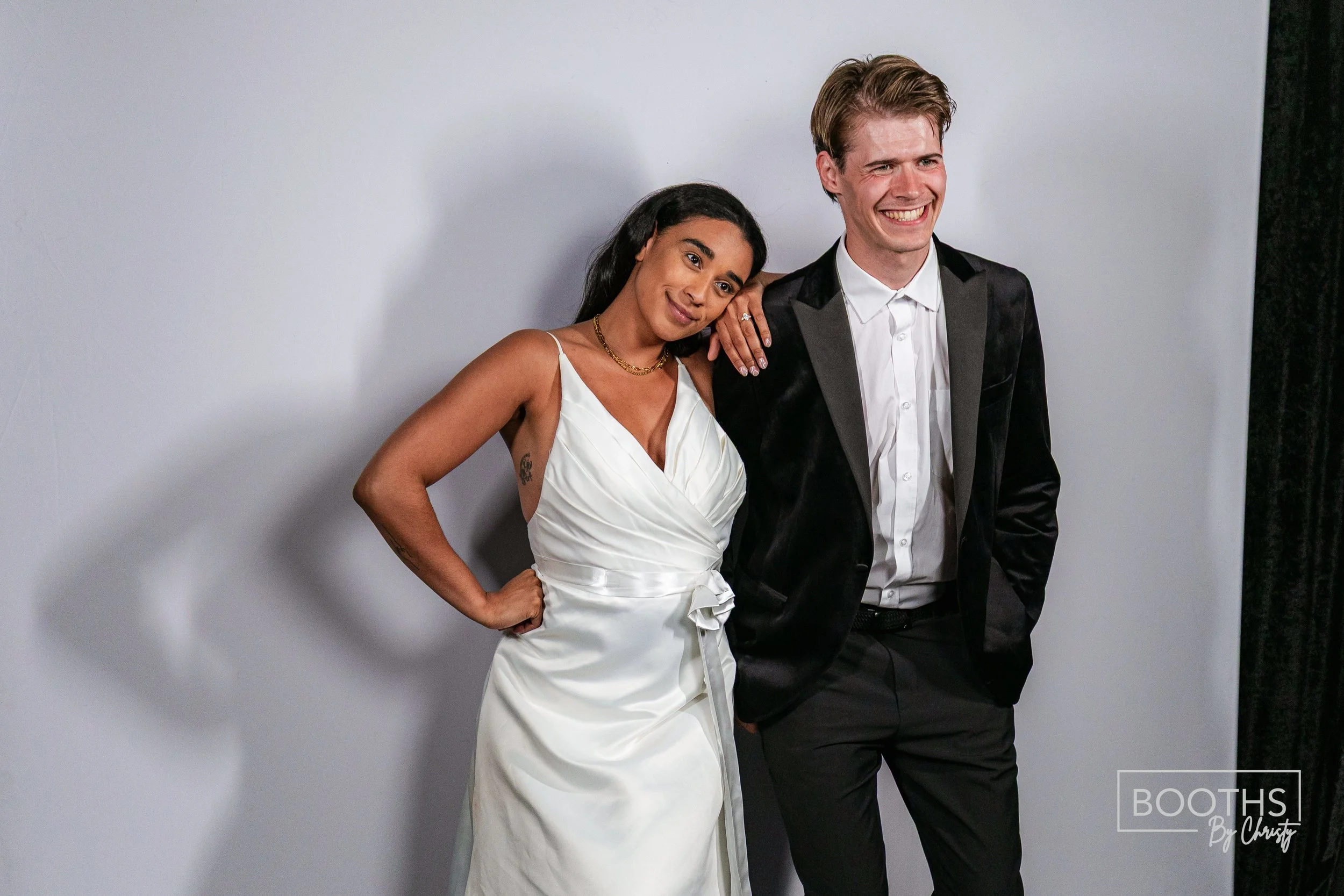 A woman in a white dress and a man in a black tuxedo pose together, smiling, against a plain background. The woman has her arm around the man's shoulder and the man has his hands in his pockets.