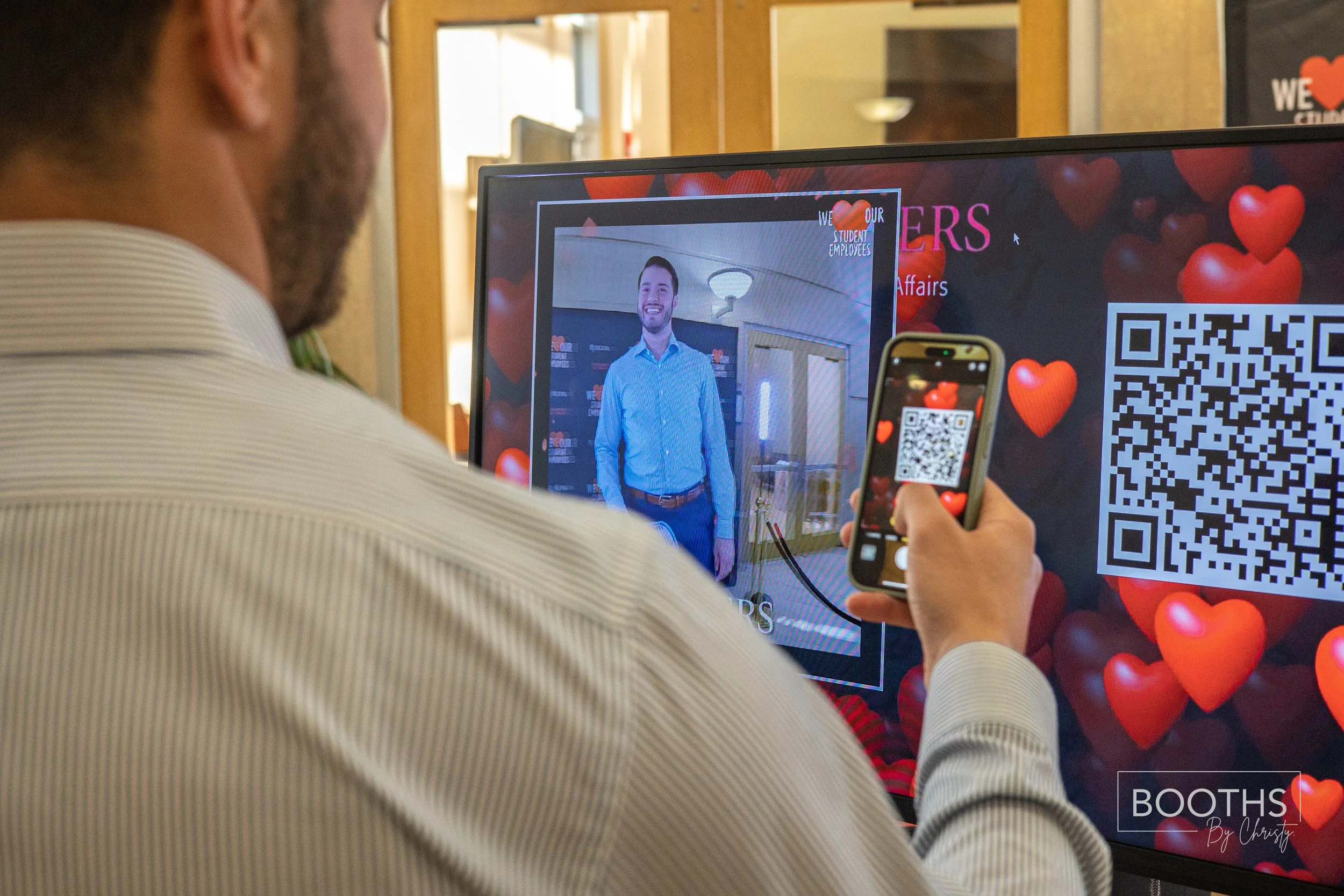 A man taking a photo of himself with his smartphone in front of a display screen with heart-shaped graphics and QR codes.