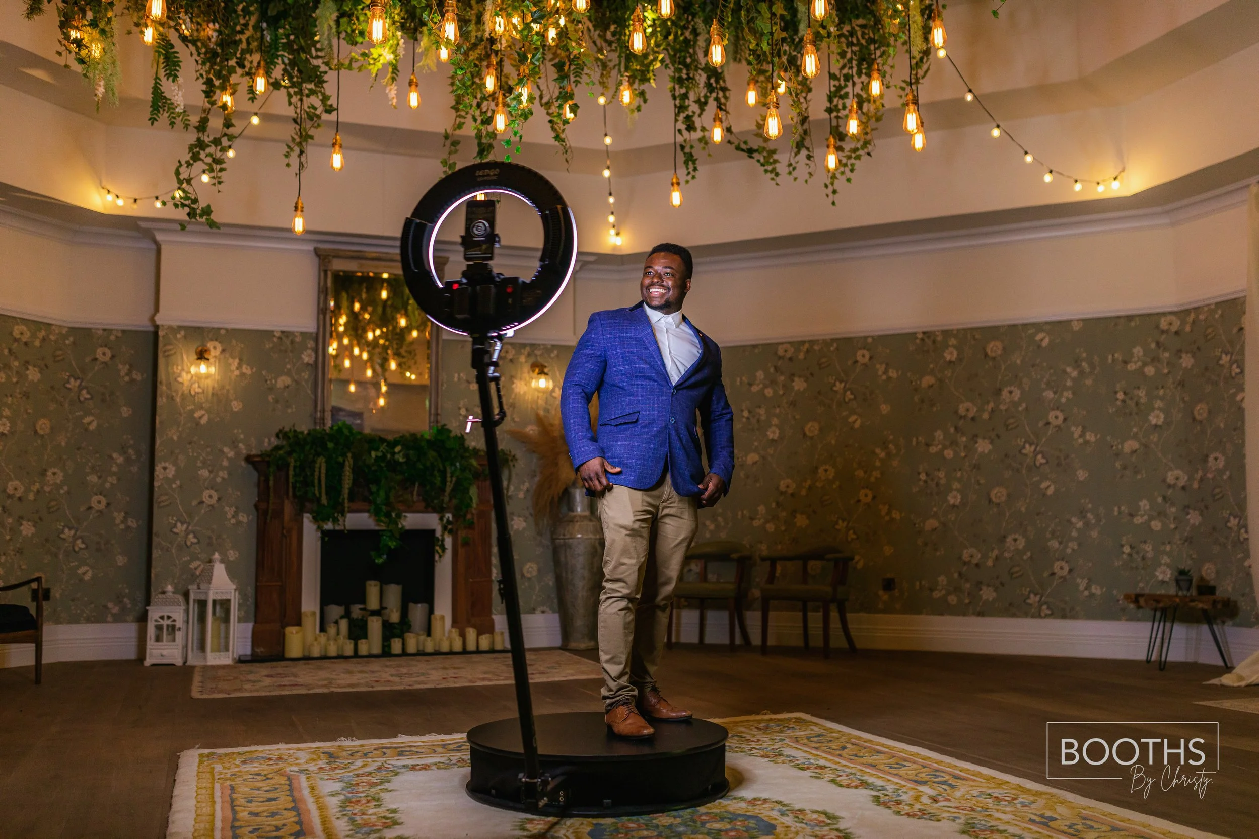 A man stands on a small circular platform in a decorated indoor event space, smiling, with a camera and lighting set up in front of him. The space has warm hanging string lights and floral wallpaper.