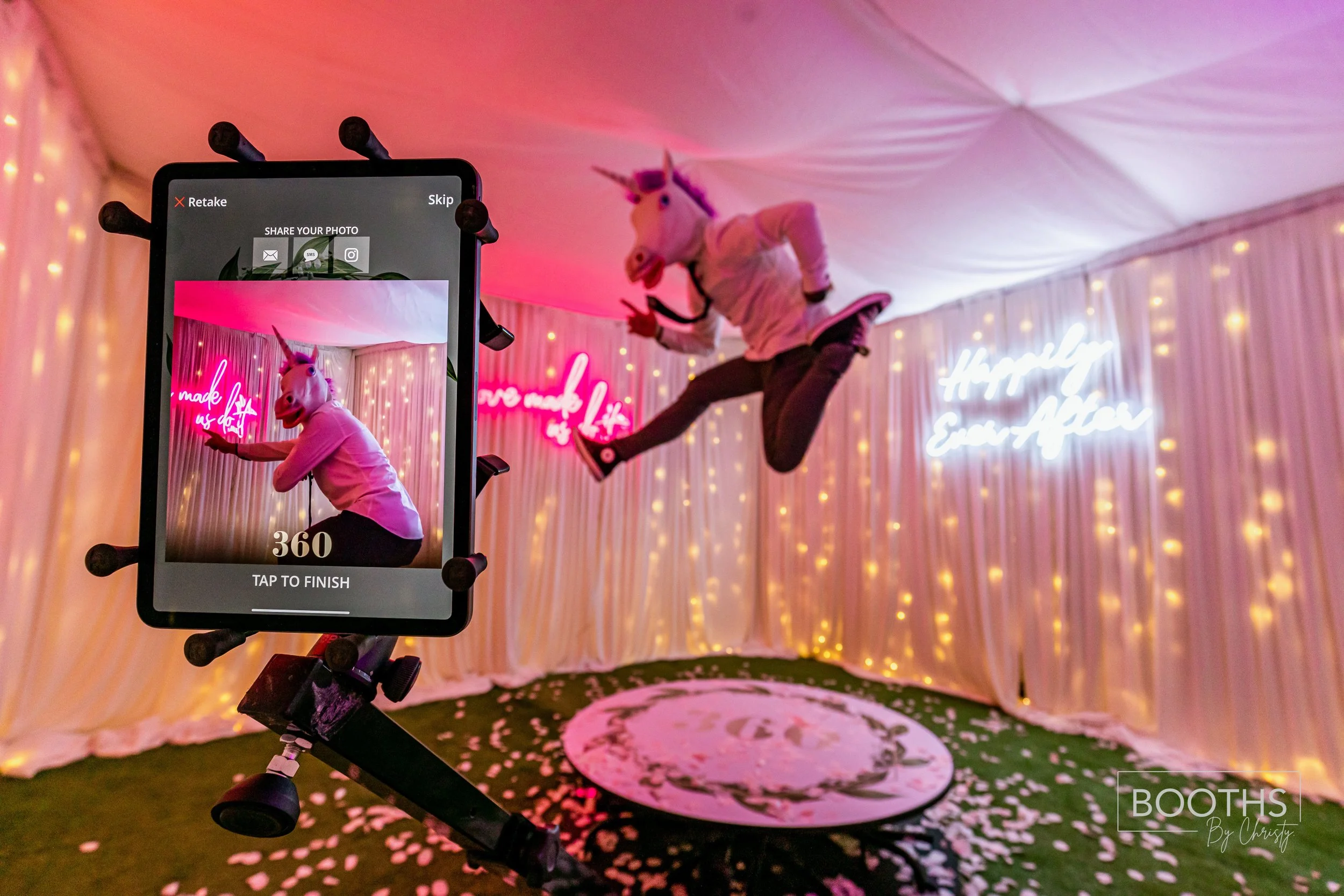 Person in a unicorn mask jumping in front of a decorated backdrop with pink neon signs and string lights, taking a selfie with a smartphone mounted on a selfie stick.