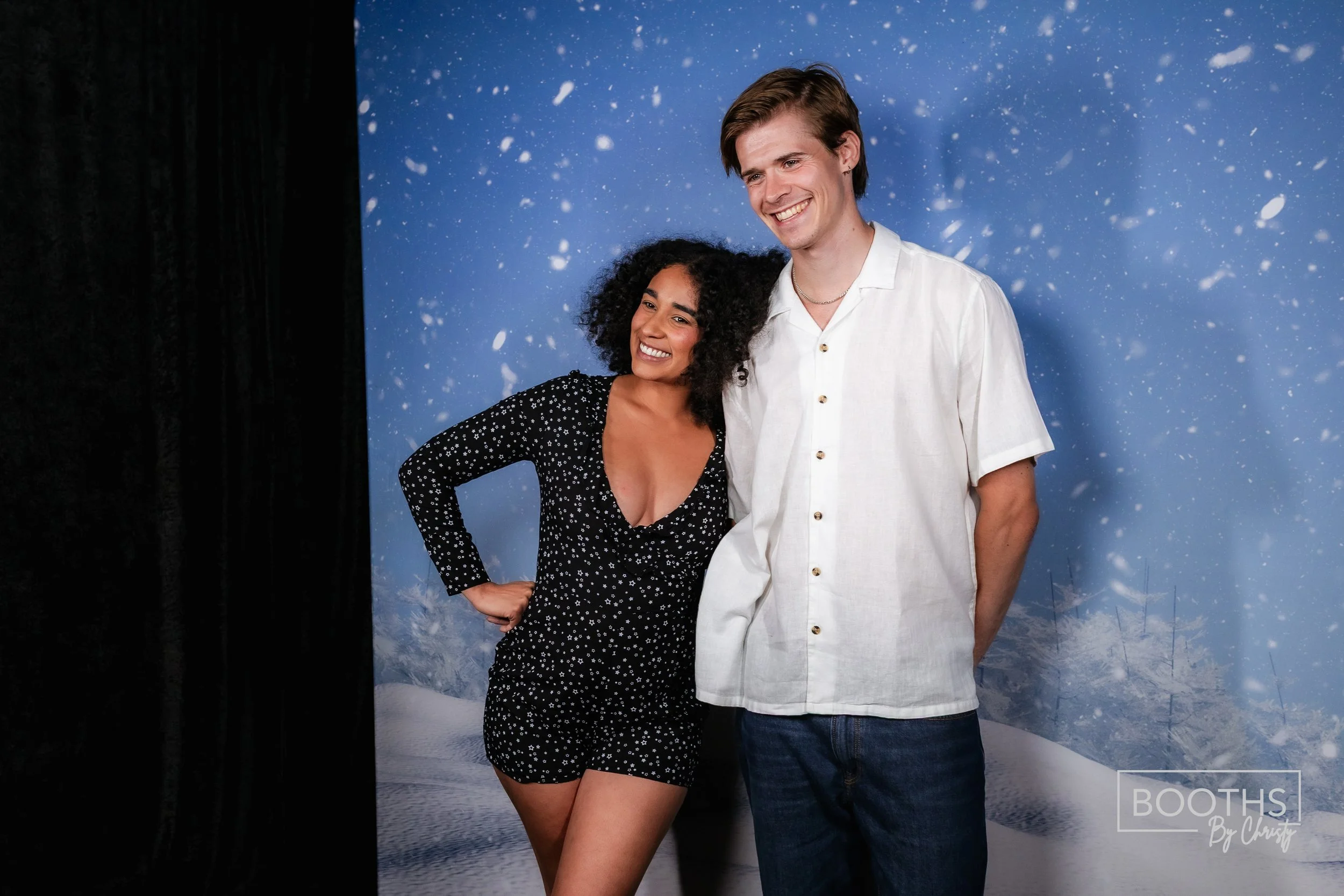A smiling woman with curly hair in a black dress with white polka dots, standing next to a smiling man with short hair in a white shirt, in front of a blue winter-themed backdrop with snowflakes.