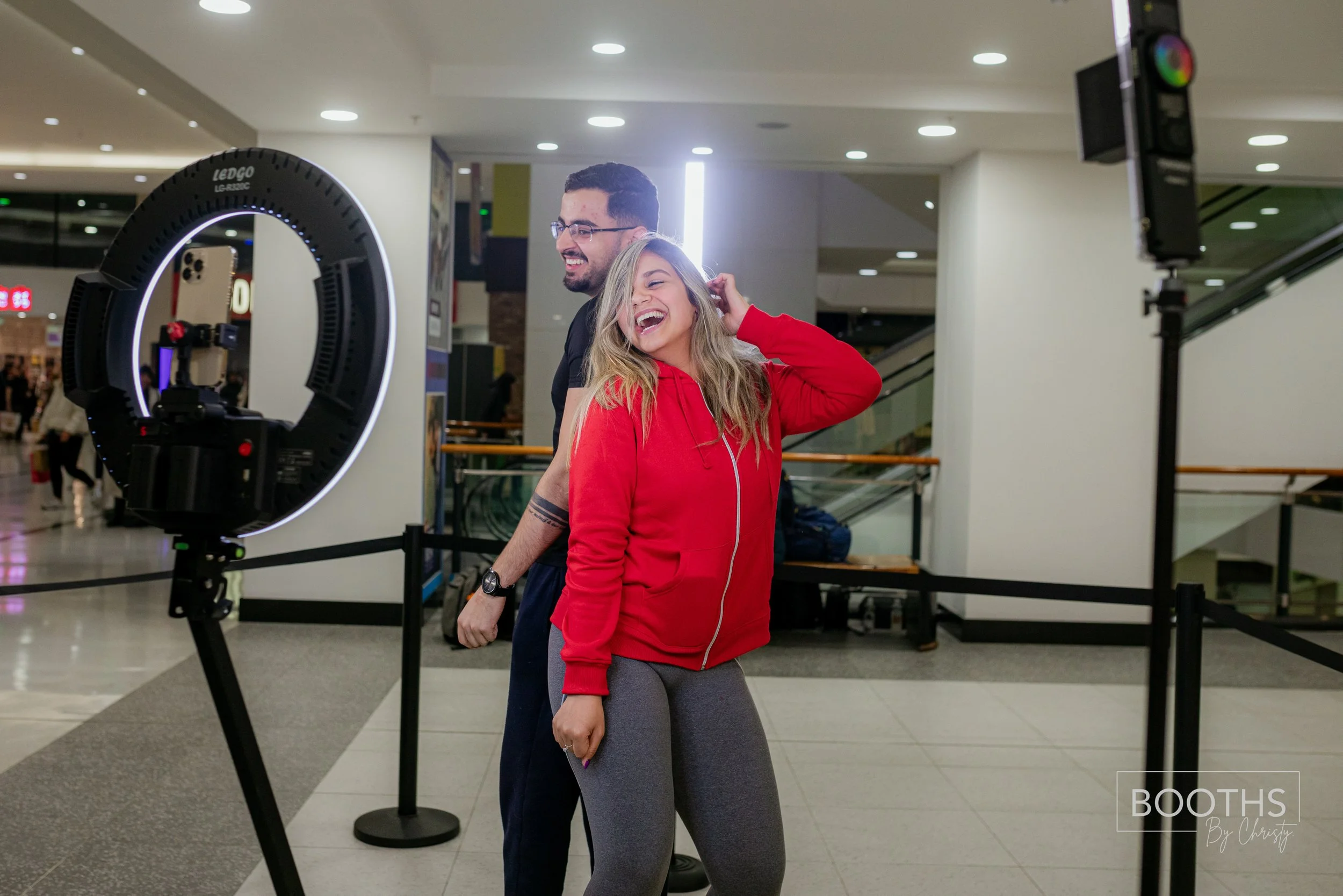 Two people standing in front of a laser ring light at an indoor location, smiling and laughing. The woman in a red hoodie and gray leggings, the man in a black shirt, with a background of an escalator and shoppers.
