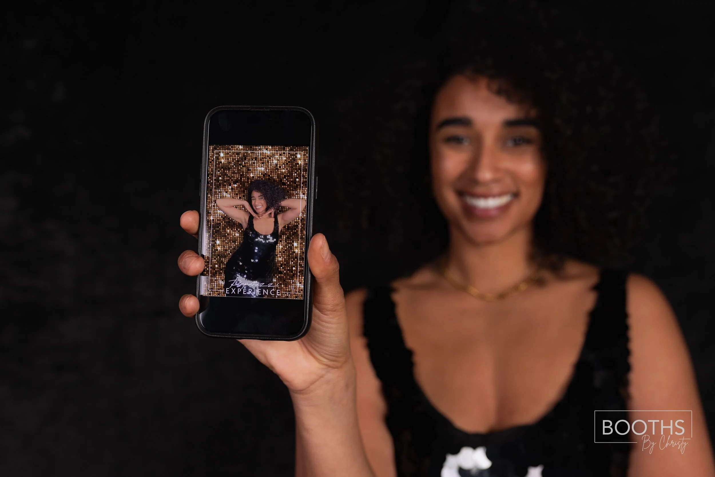 A woman with curly hair smiling while holding a phone showing her picture against a gold glittery background. The photo on the phone has the word 'Experience' on it, and the woman is wearing a black dress.