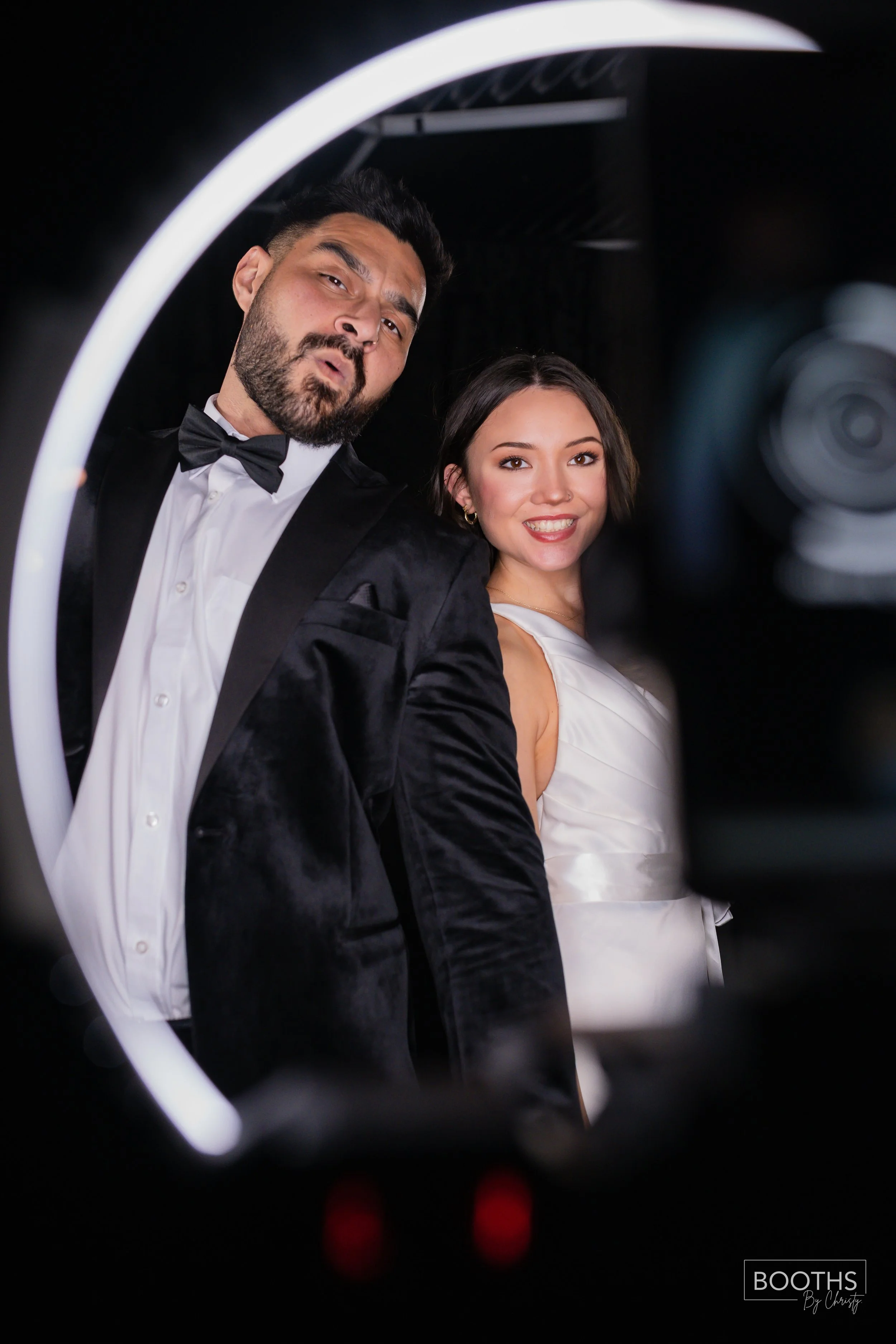 A man and woman dressed in formal attire posing for a photo, seen through a ring light and camera.