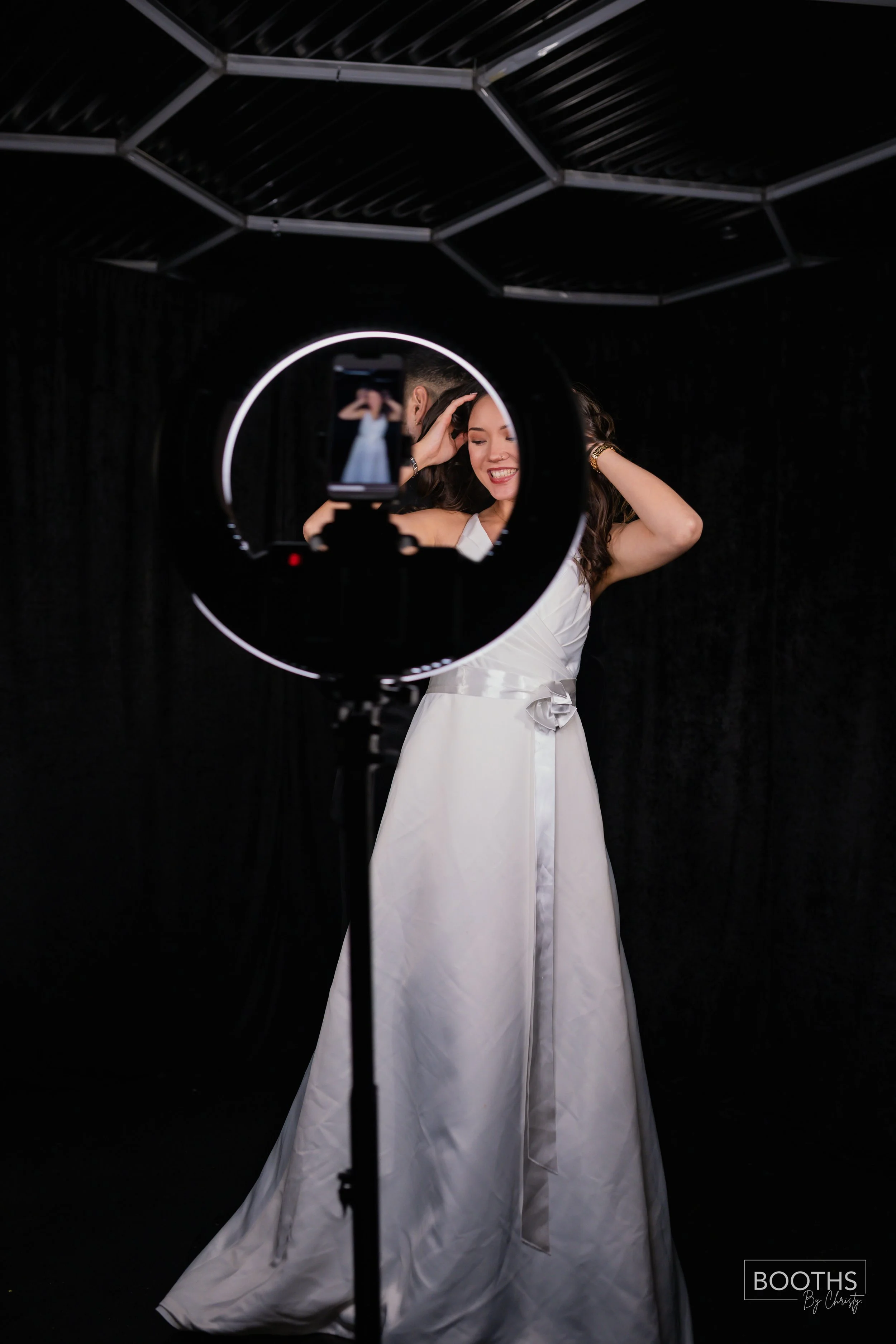 A woman in a white wedding dress is smiling and posing for a photo in front of a black backdrop, with her reflection visible in a ring light and phone camera.