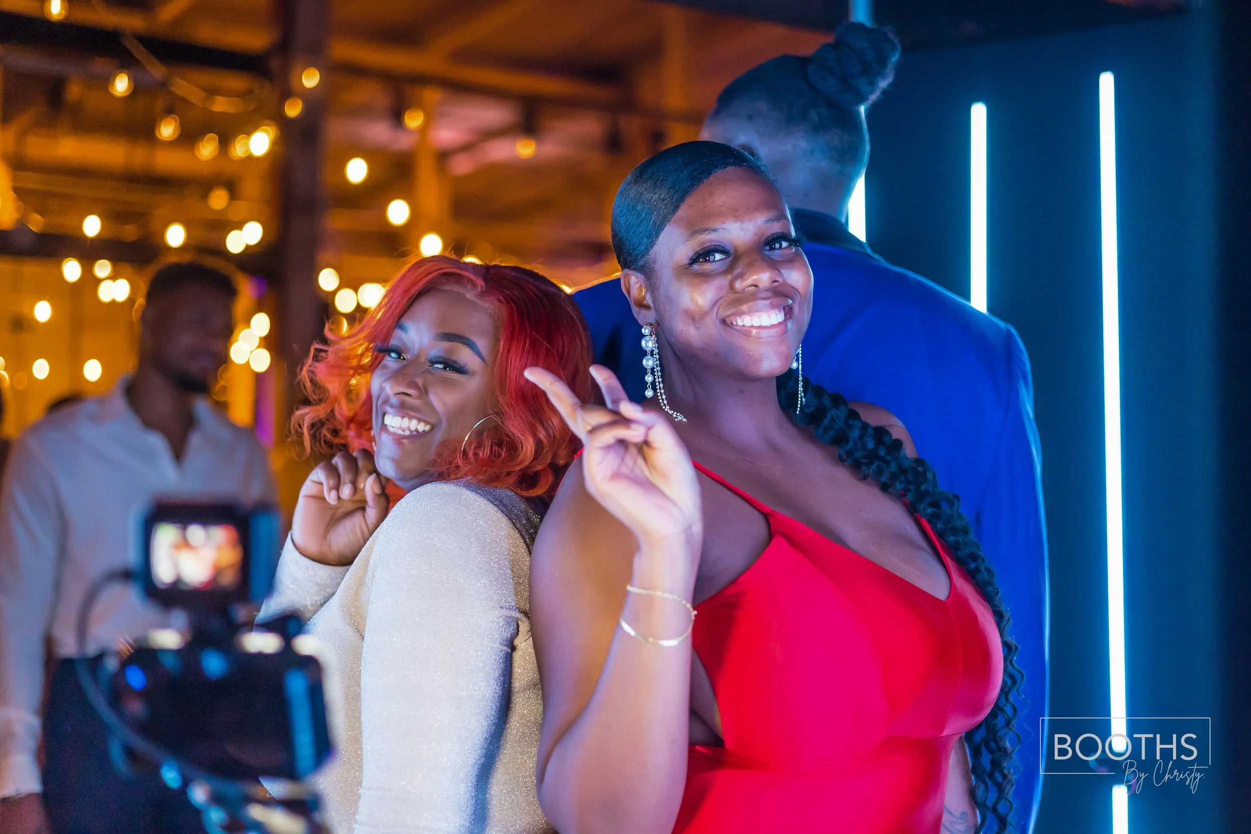 Two women smiling and posing at a party, one with red hair and the other with long braided hair, with a camera in front of them and a man in the background