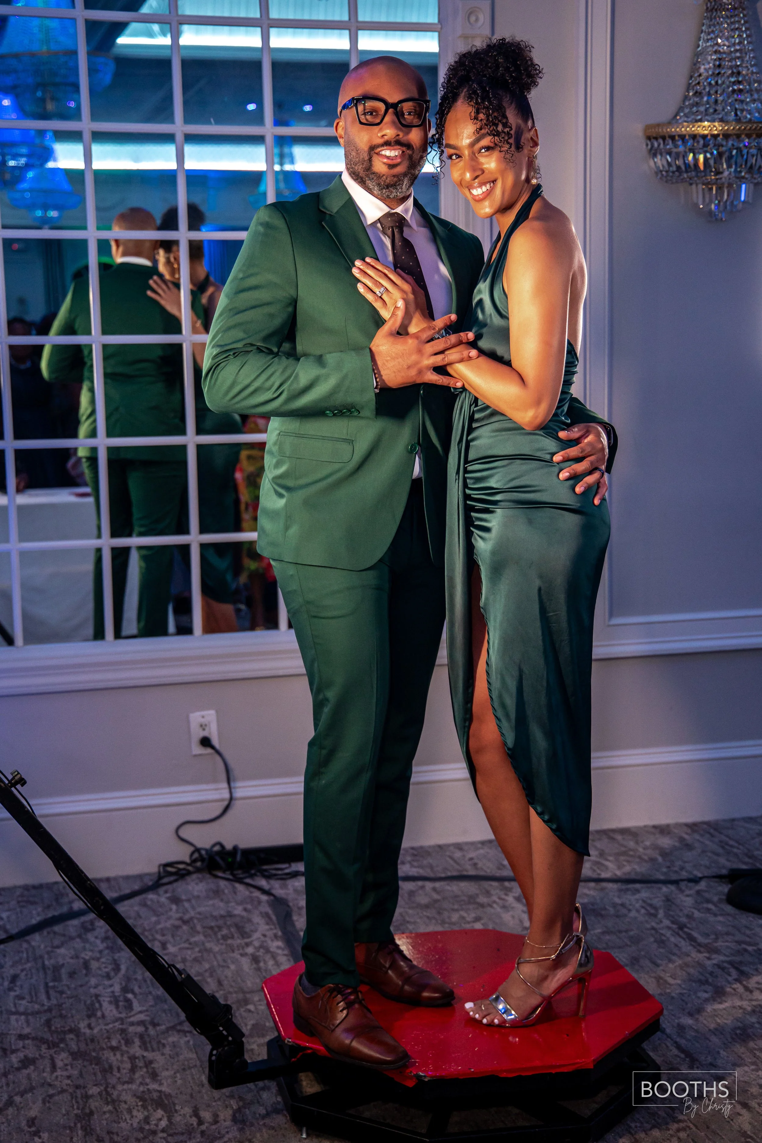 A well-dressed couple standing on a red platform at an indoor event, with a large window and a chandelier behind them. The man wears a green suit, white shirt, and black tie, and the woman wears a dark green satin dress with a high slit. They are smi