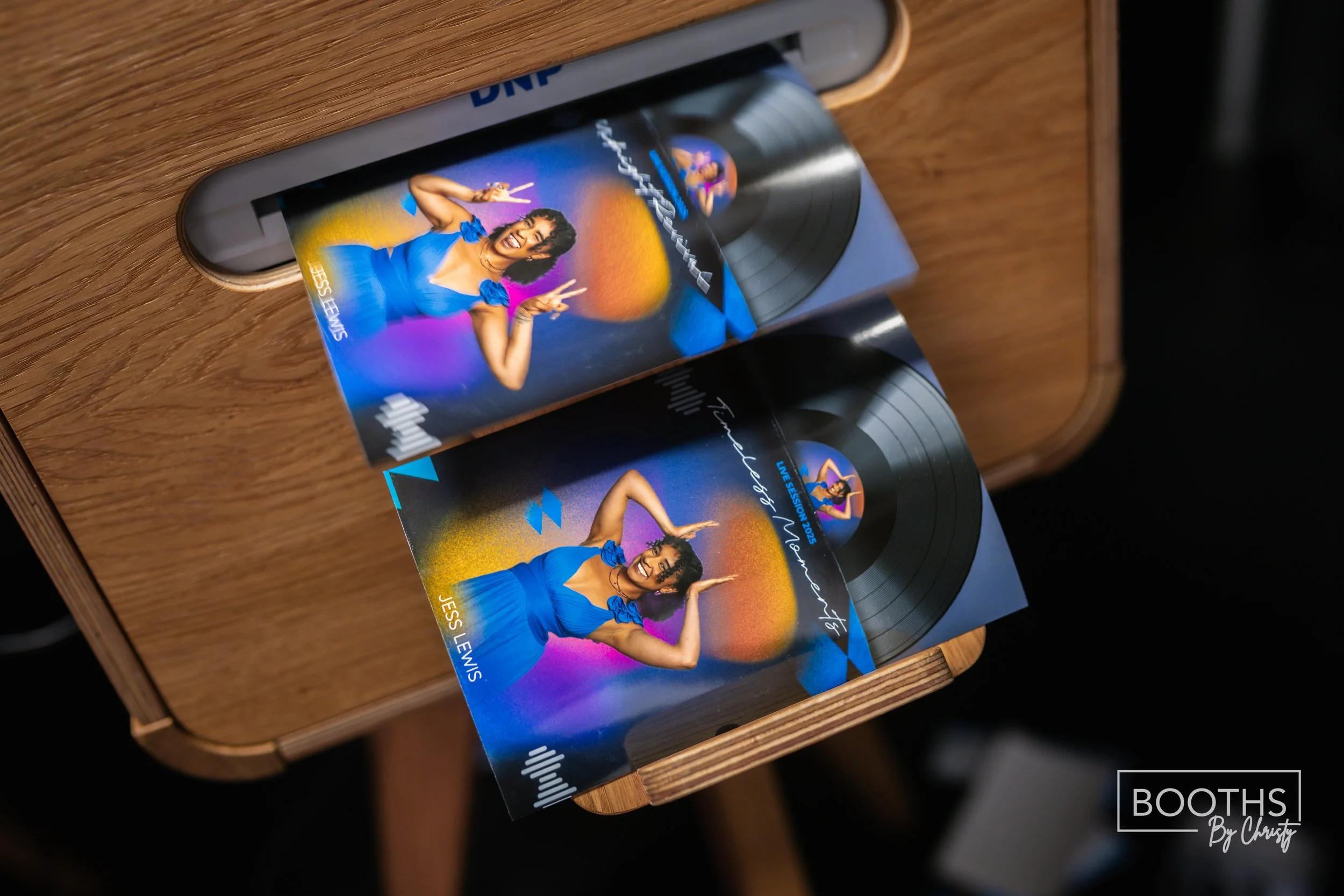 Promotional flyers for Jessi Lewis' live session 2023, featuring a woman in a blue dress making peace signs in front of colorful, abstract background with an orange sun. The flyers are placed on a wooden table, with a vinyl record partially visible.