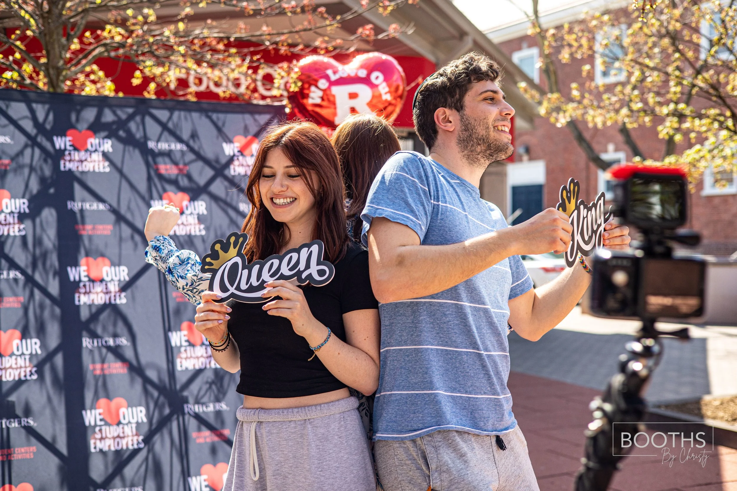 A young woman and man standing back-to-back outside at a campus event, holding signs that say "Queen" and "King". They are smiling and appear happy, with a backdrop that says "We Love Our Student Employees". There is a camera filming them and a red b