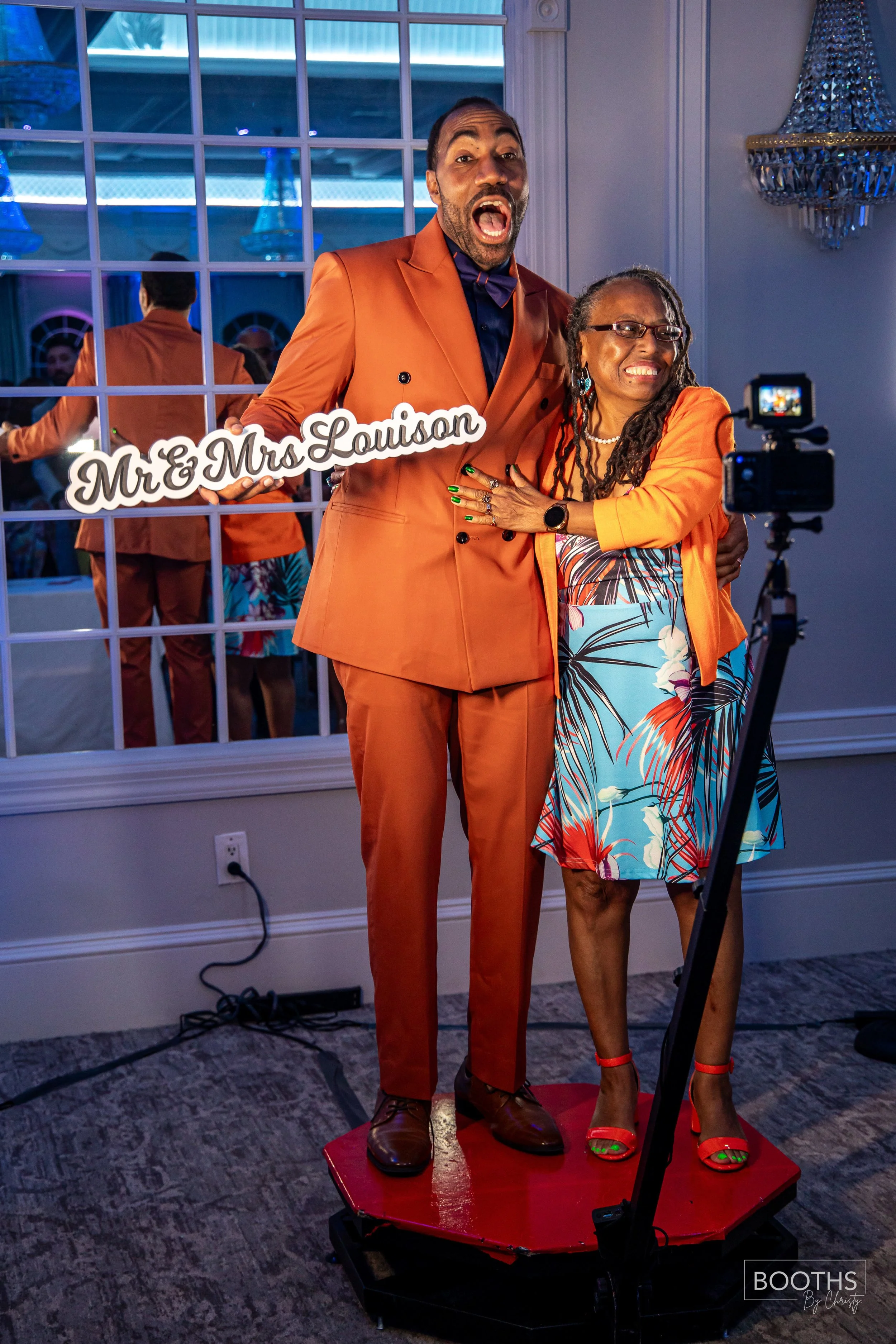 A couple dressed in vibrant orange suits taking a selfie at their wedding, holding a sign that says 'Mr. & Mrs. Louison' in a decorated indoor venue.