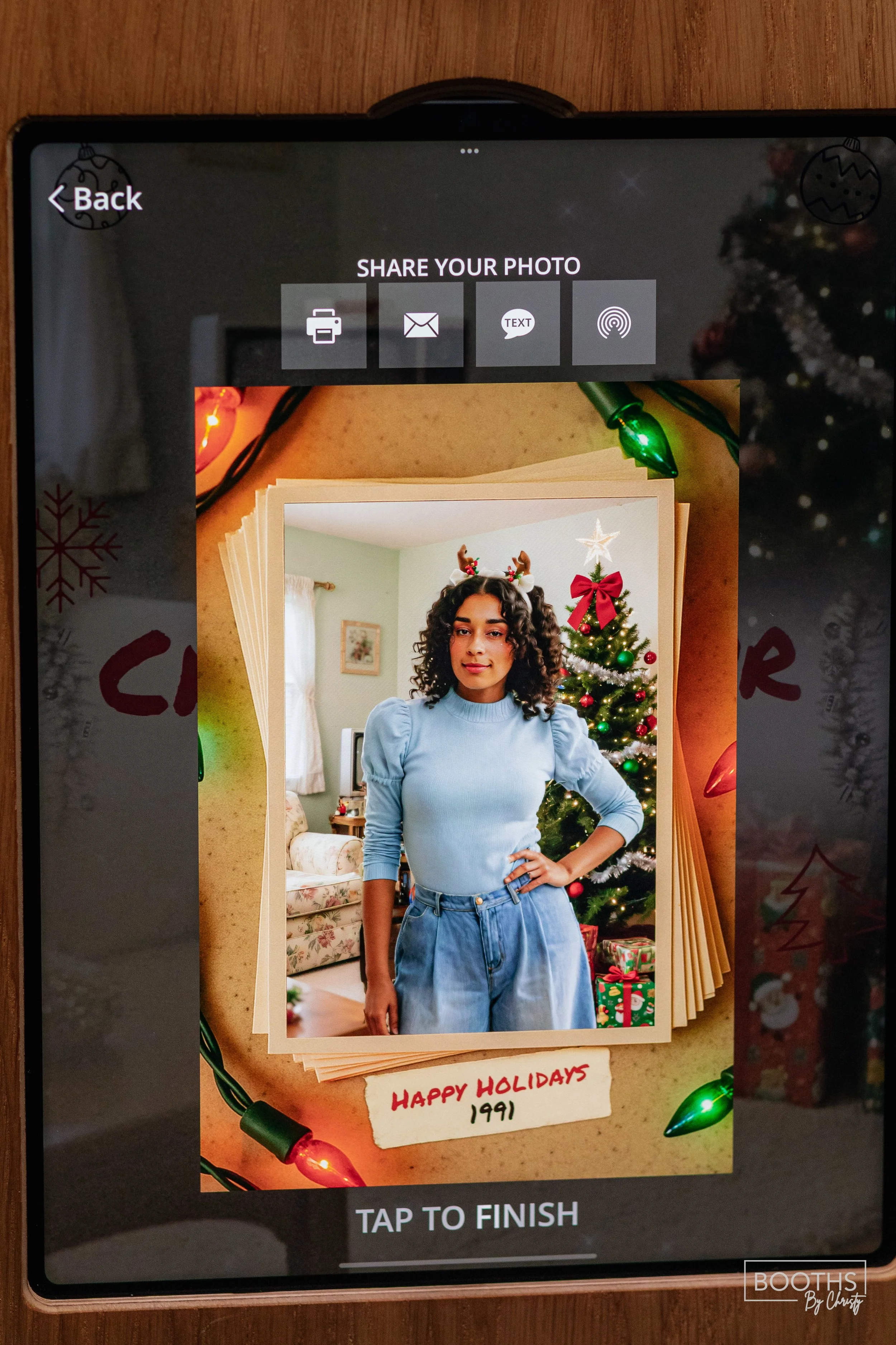 Digital photo of a woman wearing a blue sweater and jeans, with Christmas decorations in the background, including a decorated Christmas tree. The photo is displayed on a screen with a holiday-themed border and a handwritten note that reads "Happy Ho