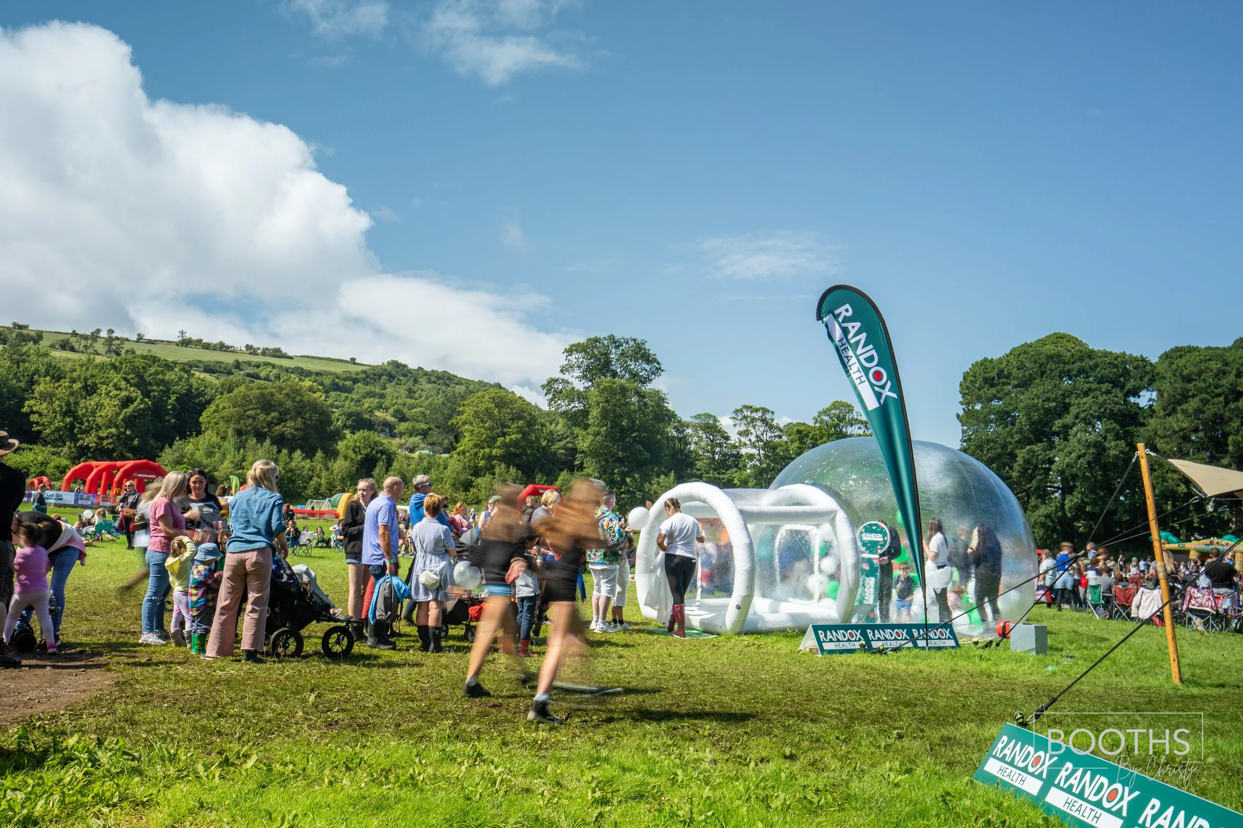 Crowd of people gathered outdoors at a health event, with a transparent inflatable pod and promotional banners for RandoX Health on a sunny day with green hills and trees in the background.