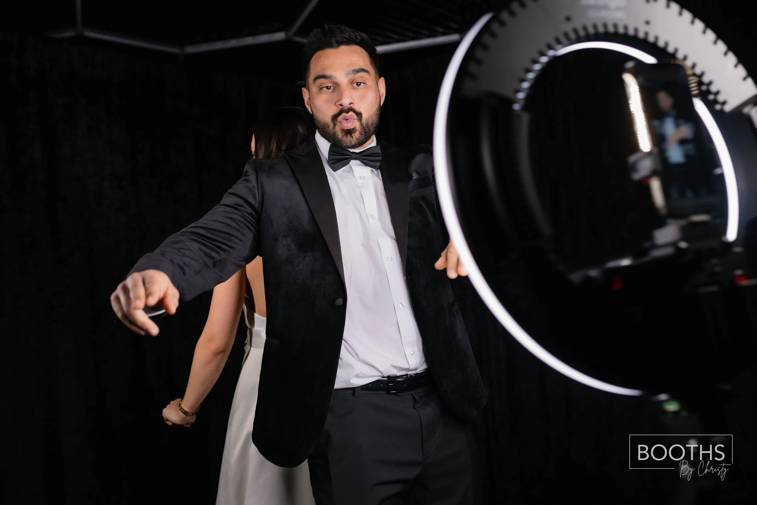 A man in a black tuxedo with a bow tie making a kissing face at a photo booth camera, with a woman in a white dress partially visible behind him, in a dark photo booth setting.