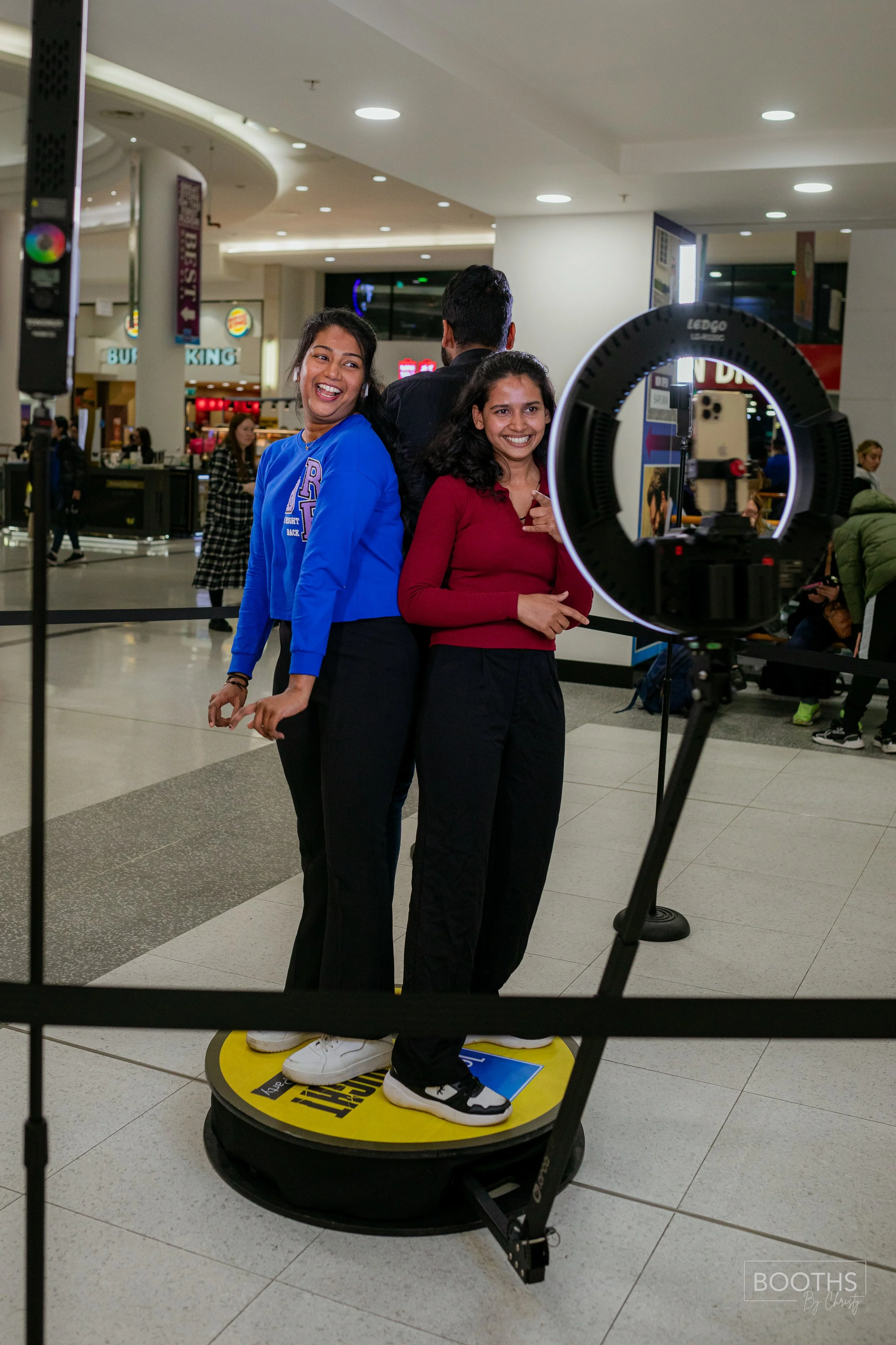 Three young women standing together for a photo in a shopping mall, with a camera setup and ring light in front of them. They are smiling and dressed casually.
