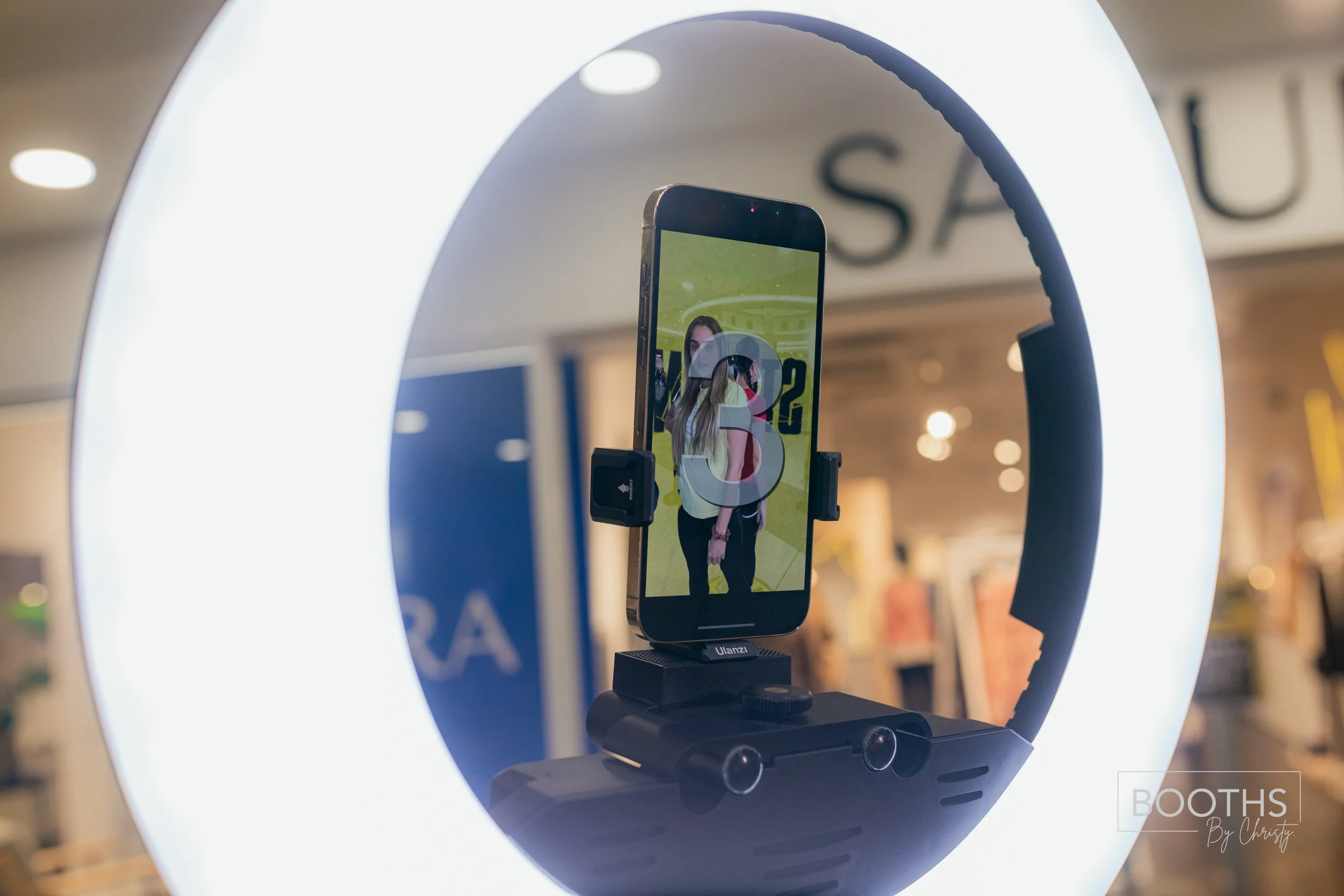 A smartphone mounted on a ring light with a woman being photographed reflected on the screen within a store.