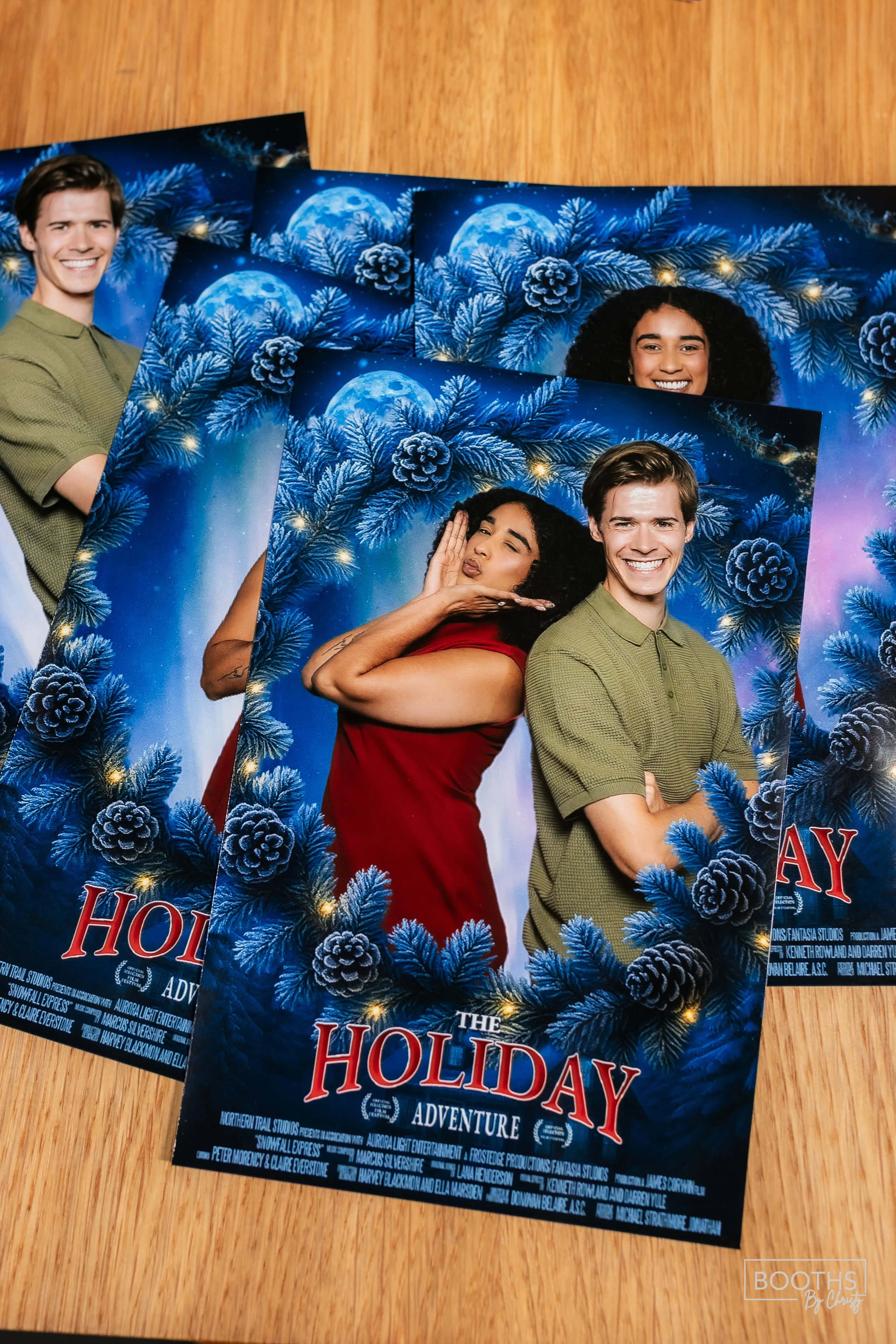 Promo photo of two smiling people, a woman with curly black hair wearing a red dress and a man with short brown hair wearing a green short-sleeved shirt, posing with a festive holiday-themed backdrop featuring pine branches, pine cones, and a full mo