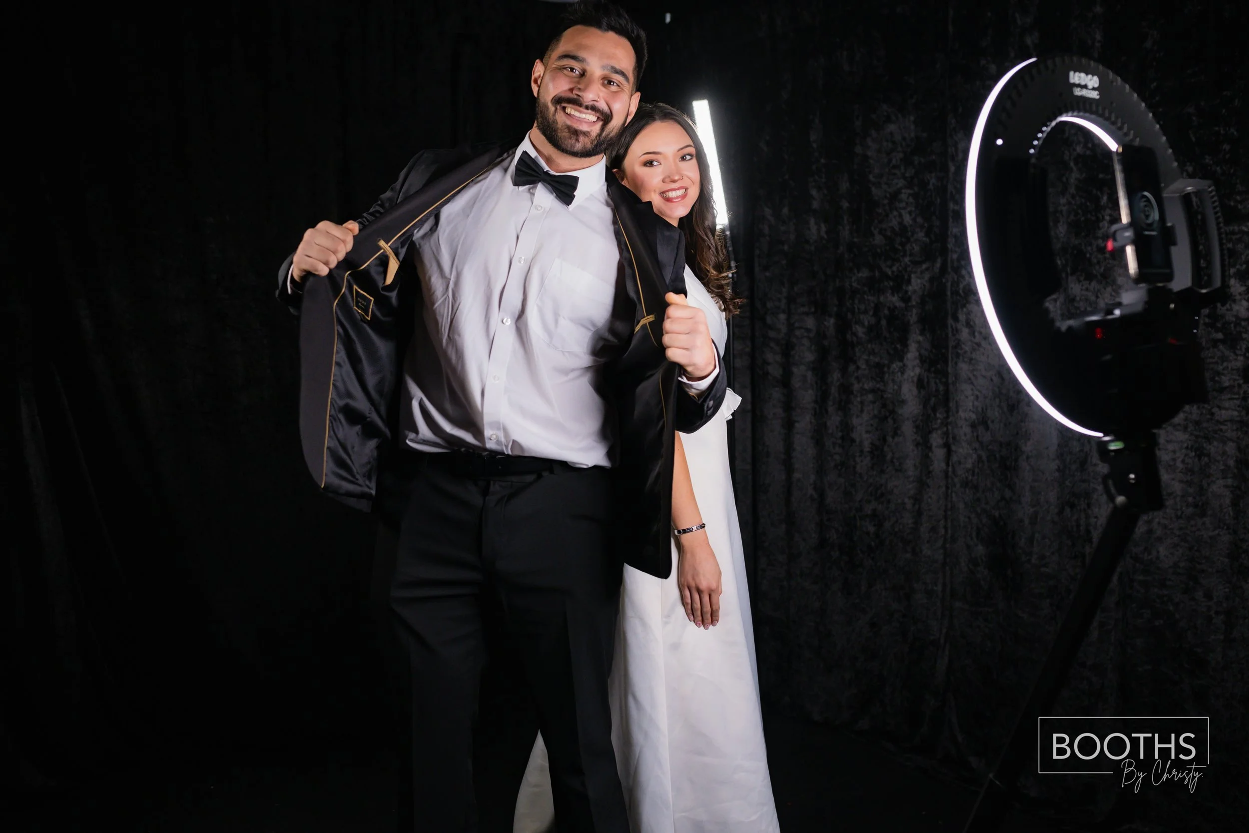 Smiling man and woman in formal attire posing for photos in a photo booth, with a camera and ring light.