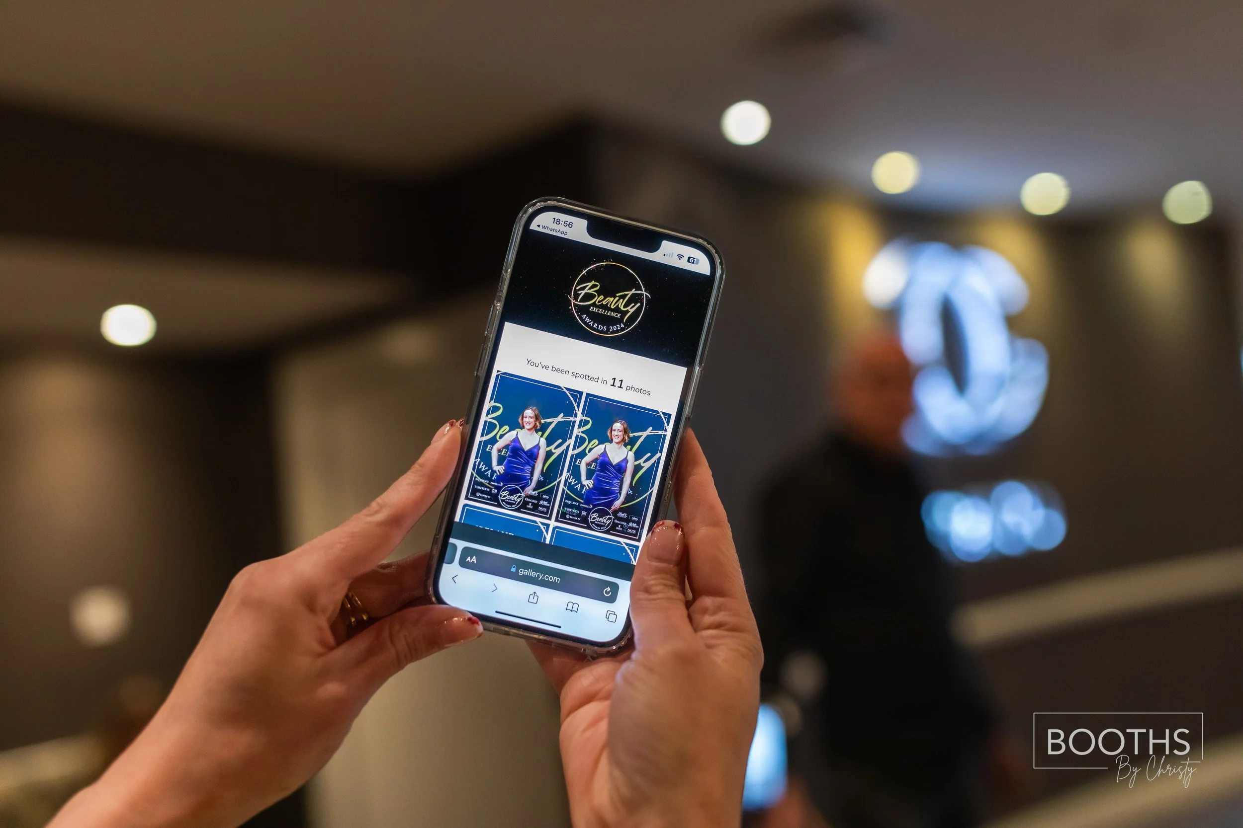 Person holding a smartphone with a photo gallery open, displaying images of an award event titled 'Beauty Excellence Awards 2024.' The background shows a blurred person and a logo on a wall.
