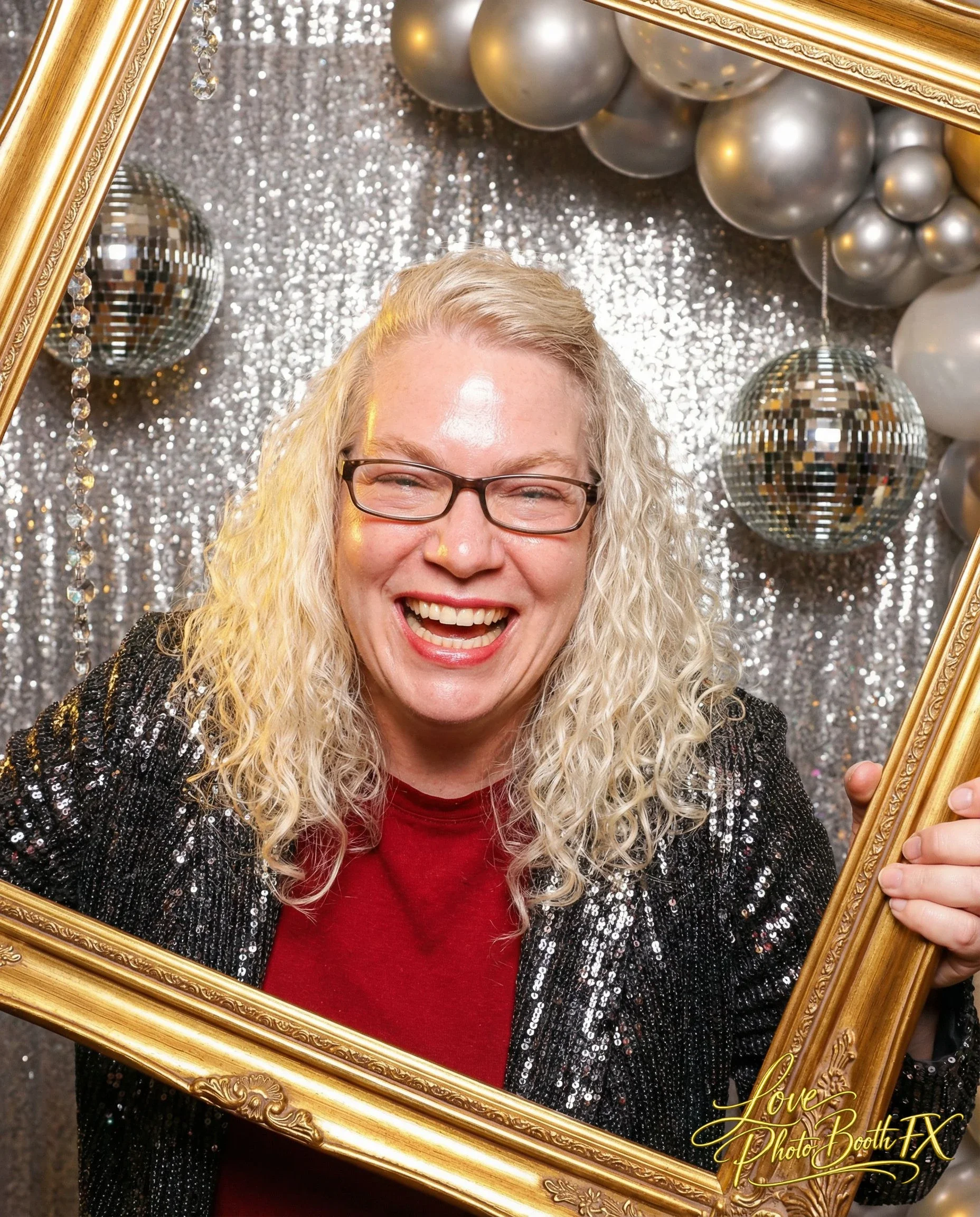 A woman with curly blonde hair, wearing glasses, a red shirt, and a black sequin jacket, smiling and holding a gold photo frame in front of a glittery silver backdrop with balloons and disco balls.