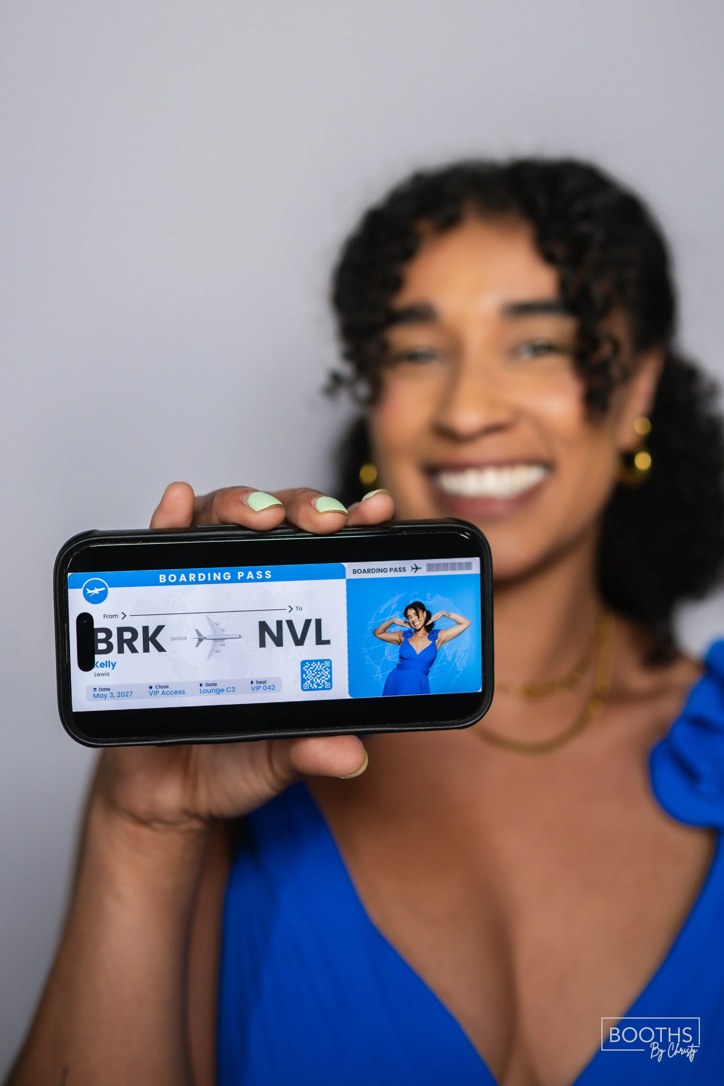 A woman in a blue dress holding up a digital boarding pass for a flight from Brooklyn (BRK) to Las Vegas (NVL), smiling, with a blurred gray background.