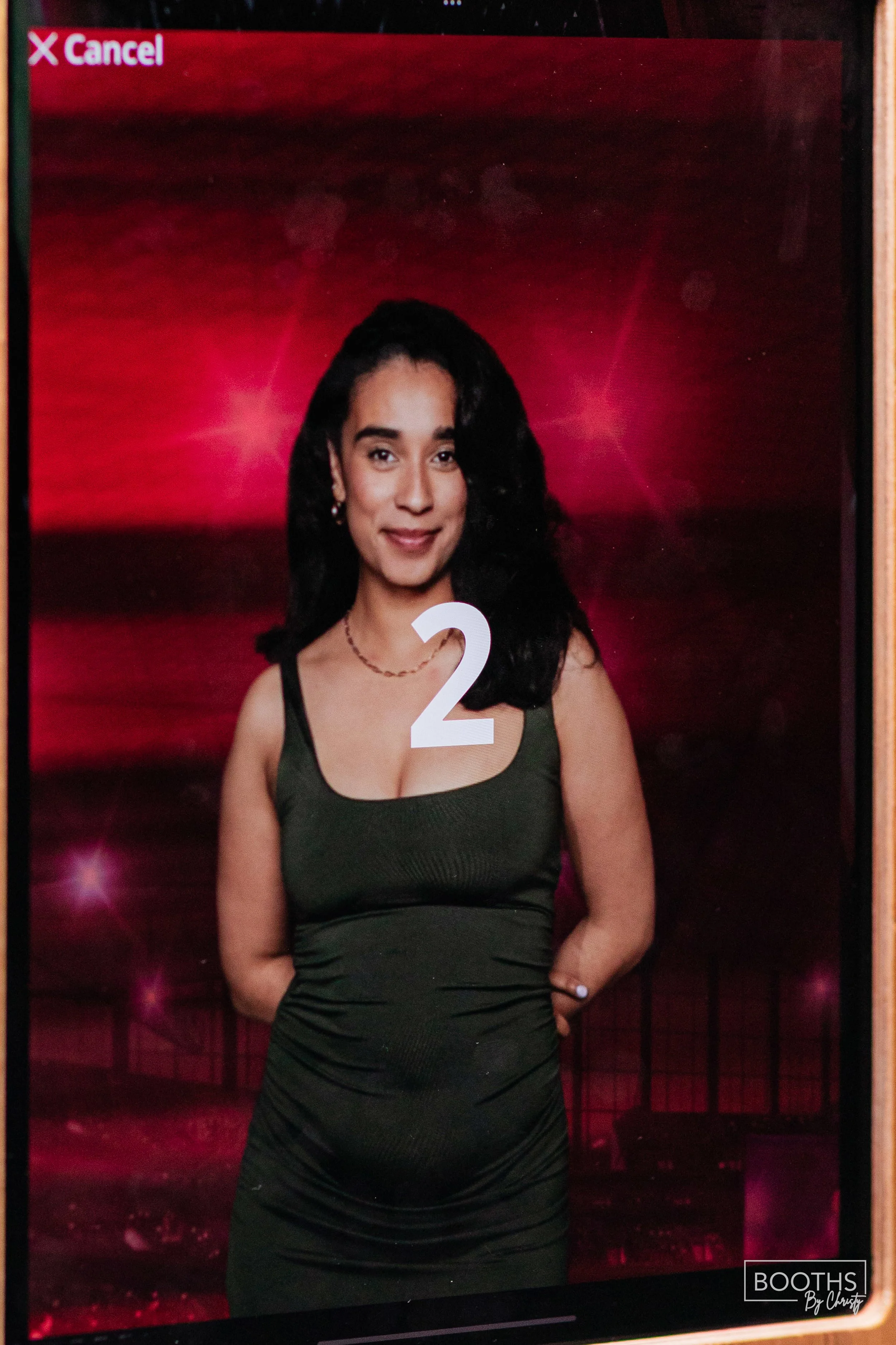 A photo booth image of a woman with dark hair and a dark green dress, standing against a red background with pink light effects. There is a large white number "2" overlayed on her chest and a red "Cancel" option in the top left corner.
