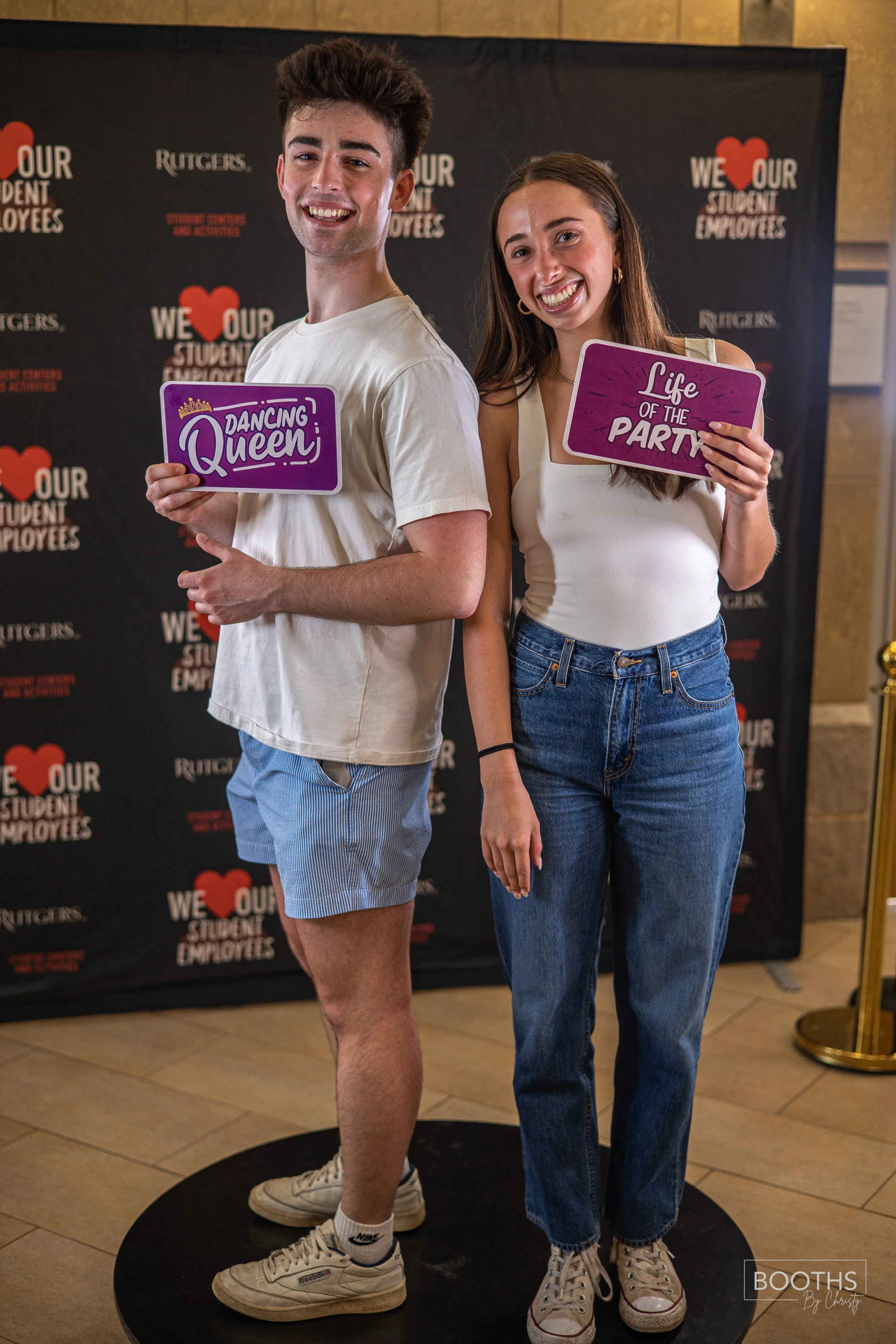 A young man and woman standing together at an event, each holding signs that say 'Dancing Queen' and 'Life of the Party.' The background features a black step-and-repeat banner with red and white text, including 'We Love Our Student Employees' and th
