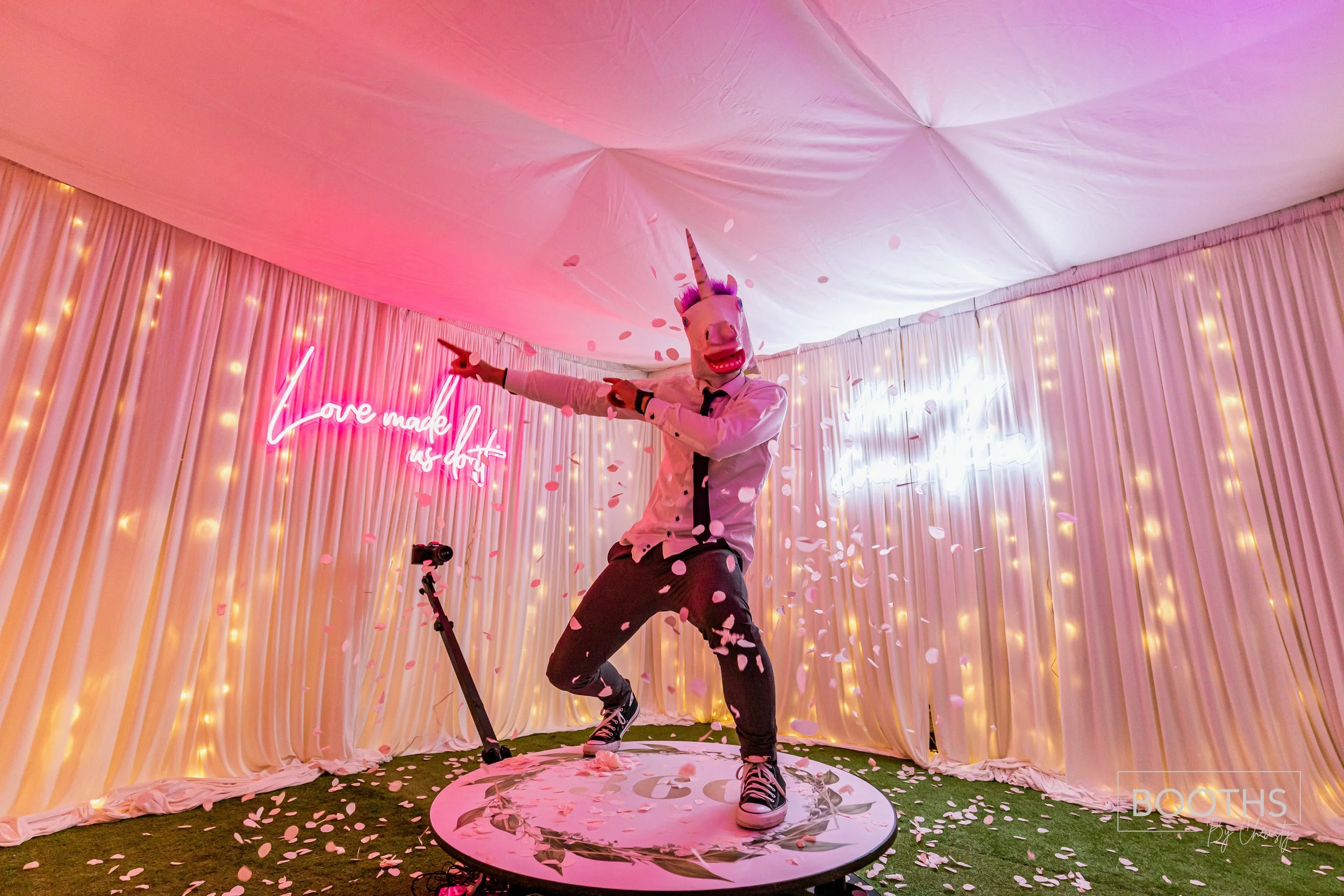 Person wearing a unicorn mask, white shirt, and dark pants dancing on a round platform with pink and white flower petals, in front of a decorated backdrop with pink and white curtains, fairy lights, and neon signs reading 'Love made us do it' and oth